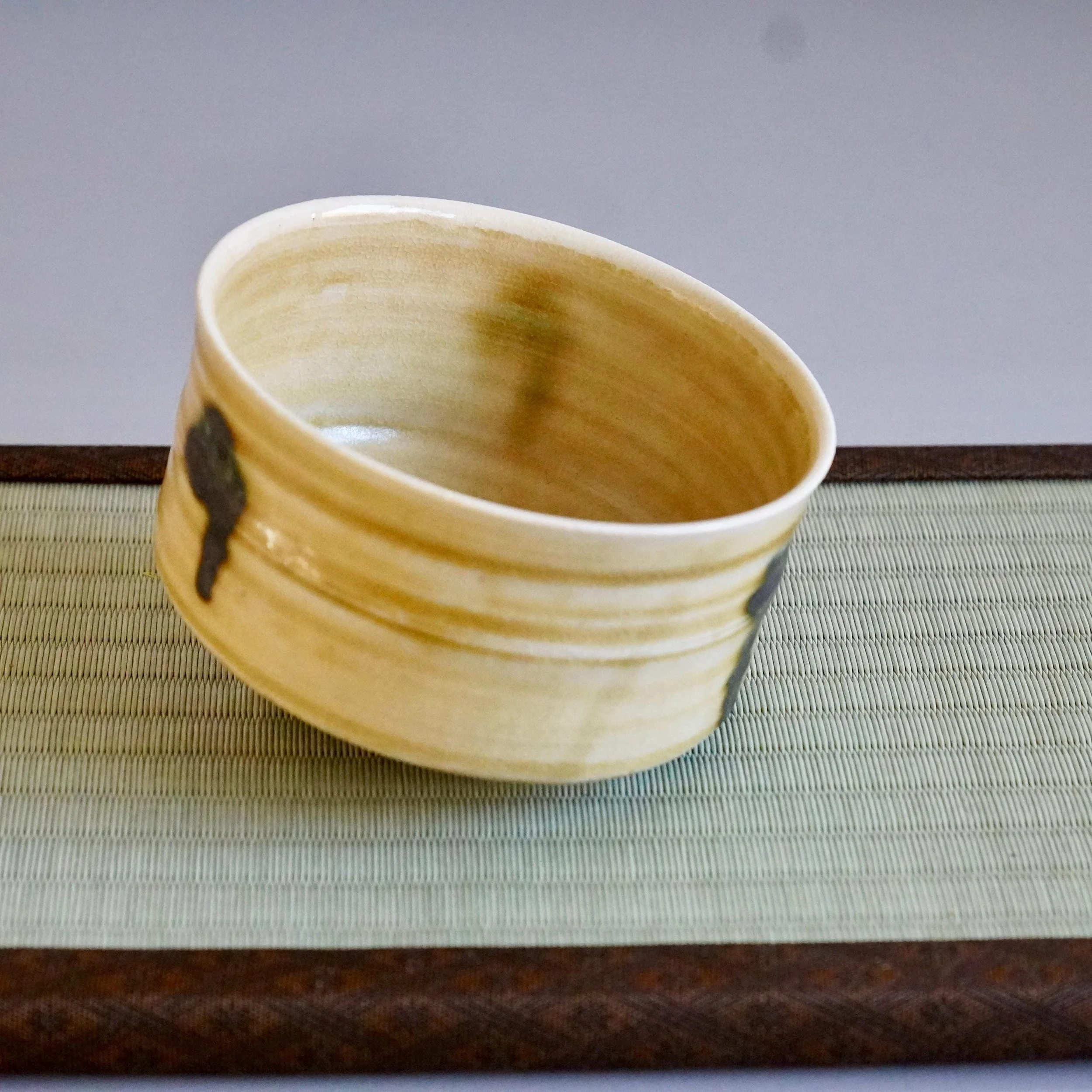 Ki-Seto chawan by Yasuda Masahiro. Low, mustard-yellow bowl with horizontal ridges, dark abstract marks, and green-brown interior washes, on a woven mat.