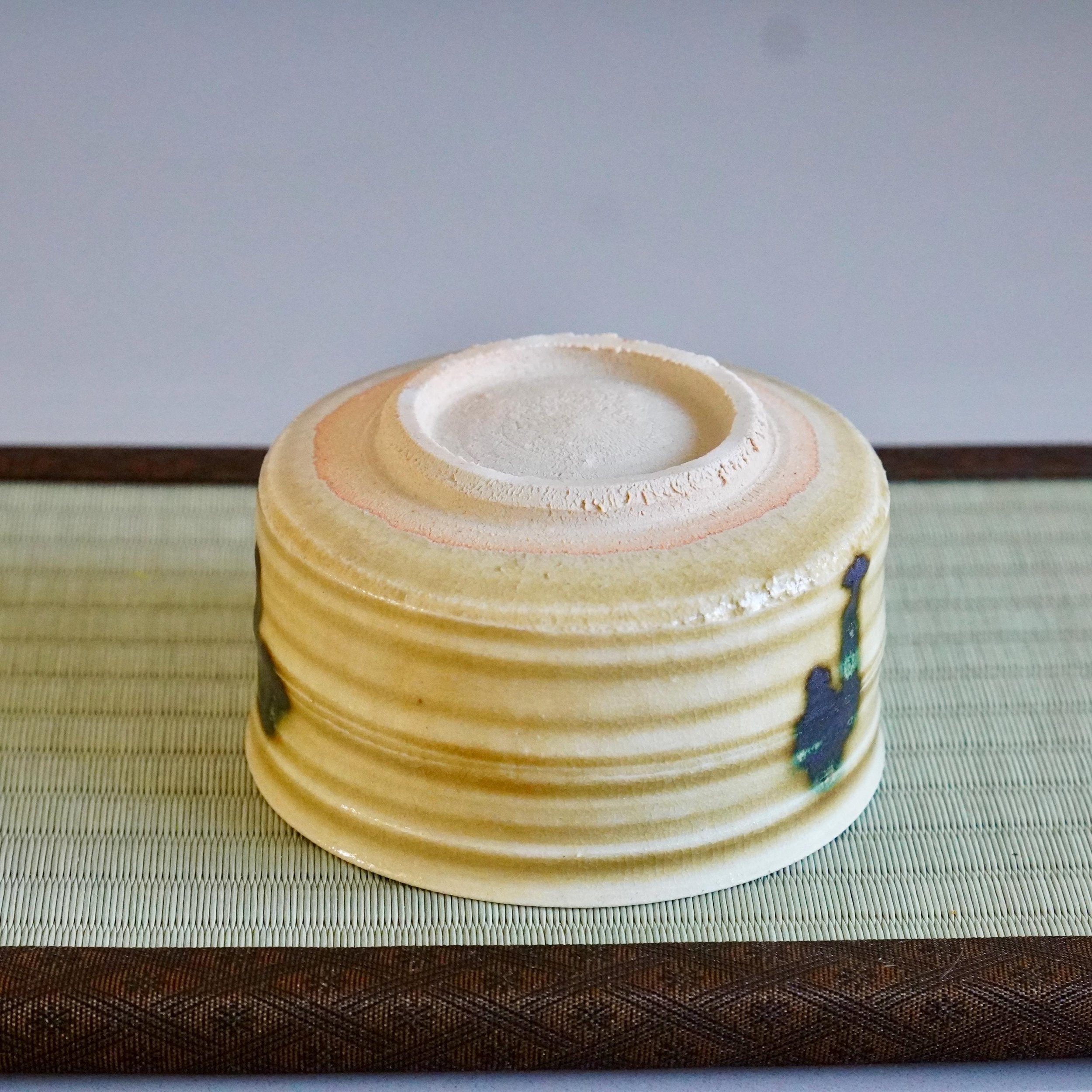 Close-up of the underside of a Ki-Seto chawan by Yasuda Masahiro, showing its mustard-yellow ridged body with blue-green glaze drips, resting on a tatami mat.