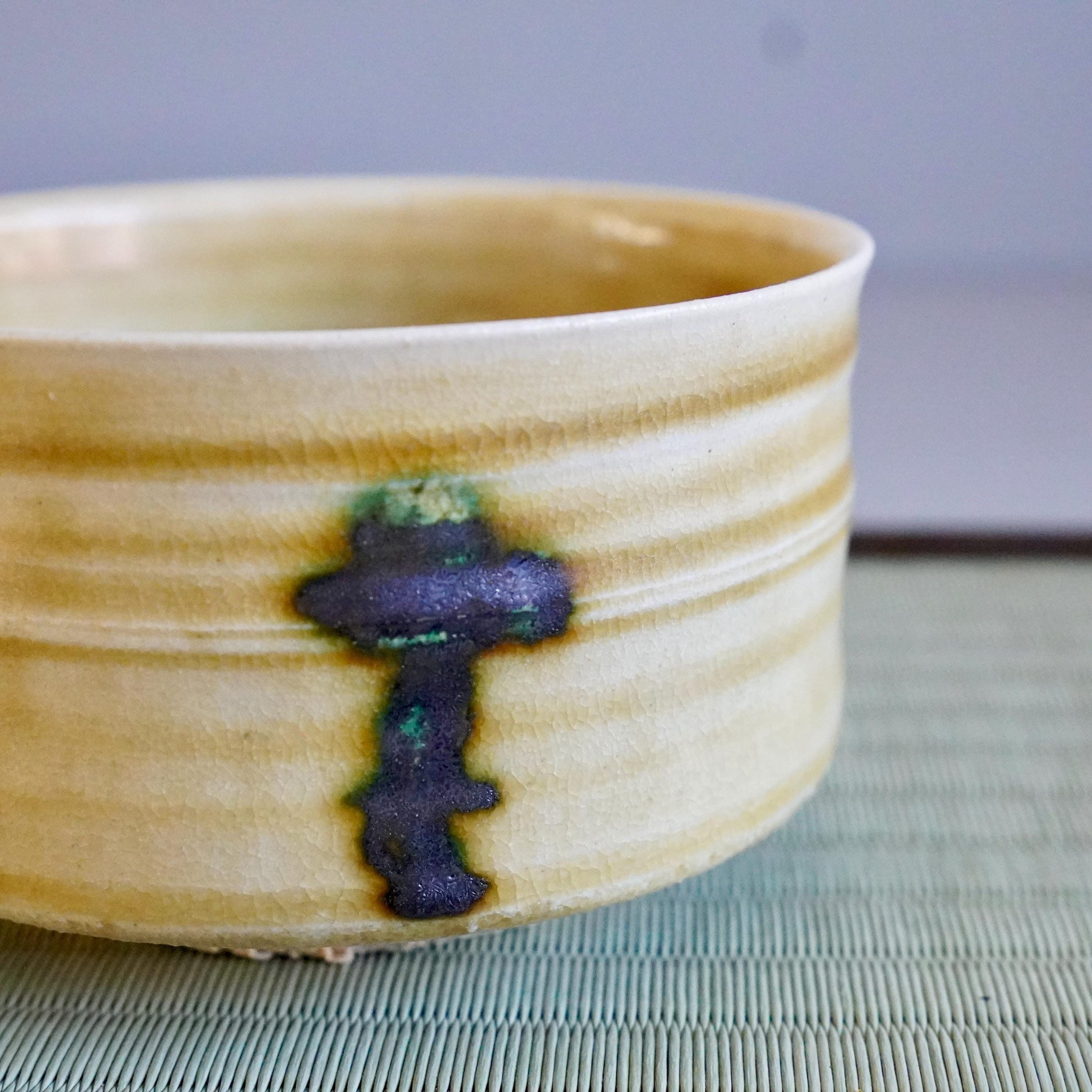 Yasuda Masahiro's mustard-yellow Ki-Seto chawan, with horizontal ridges, crackle glaze, and dark blue-green drip decoration, rests on a woven mat.