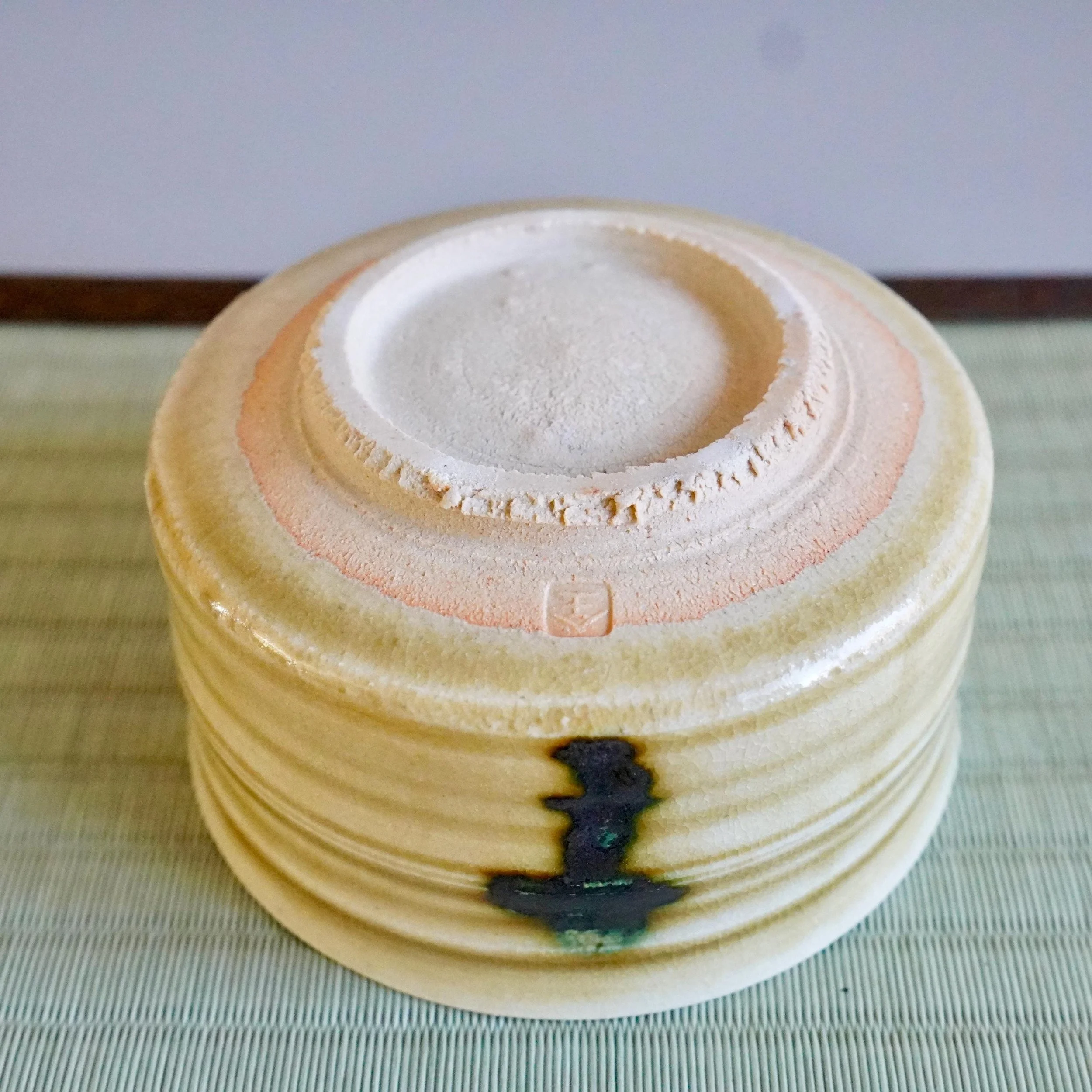 Underside of a mustard-yellow ceramic bowl by Yasuda Masahiro, its ribbed body showing a dark green glaze drip. The unglazed footring features an impressed seal, all on a woven mat.