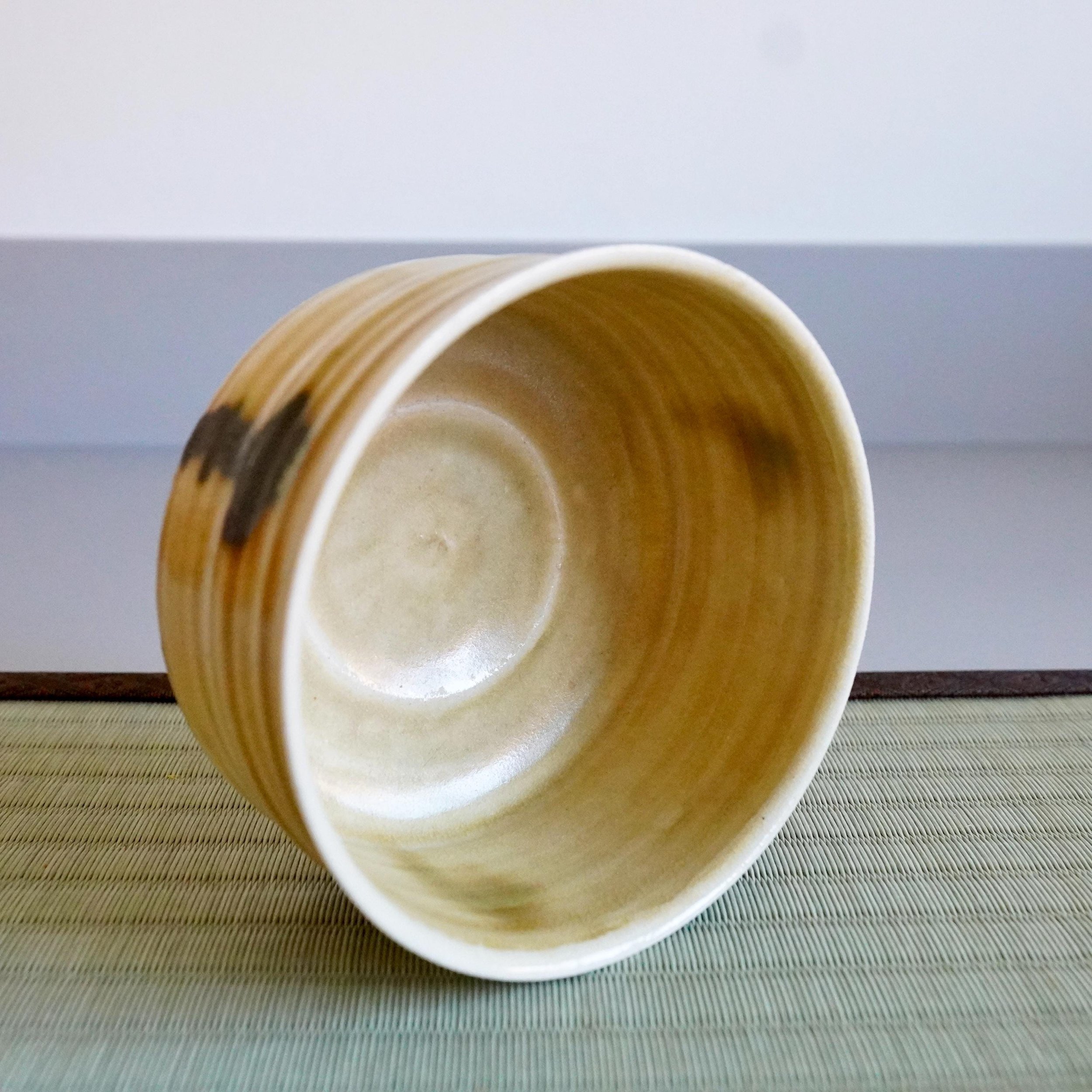 Yasuda Masahiro's mustard-yellow chawan, tilted to show its ridged exterior with dark brown marks and lighter interior with concentric circles, rests on a woven mat.