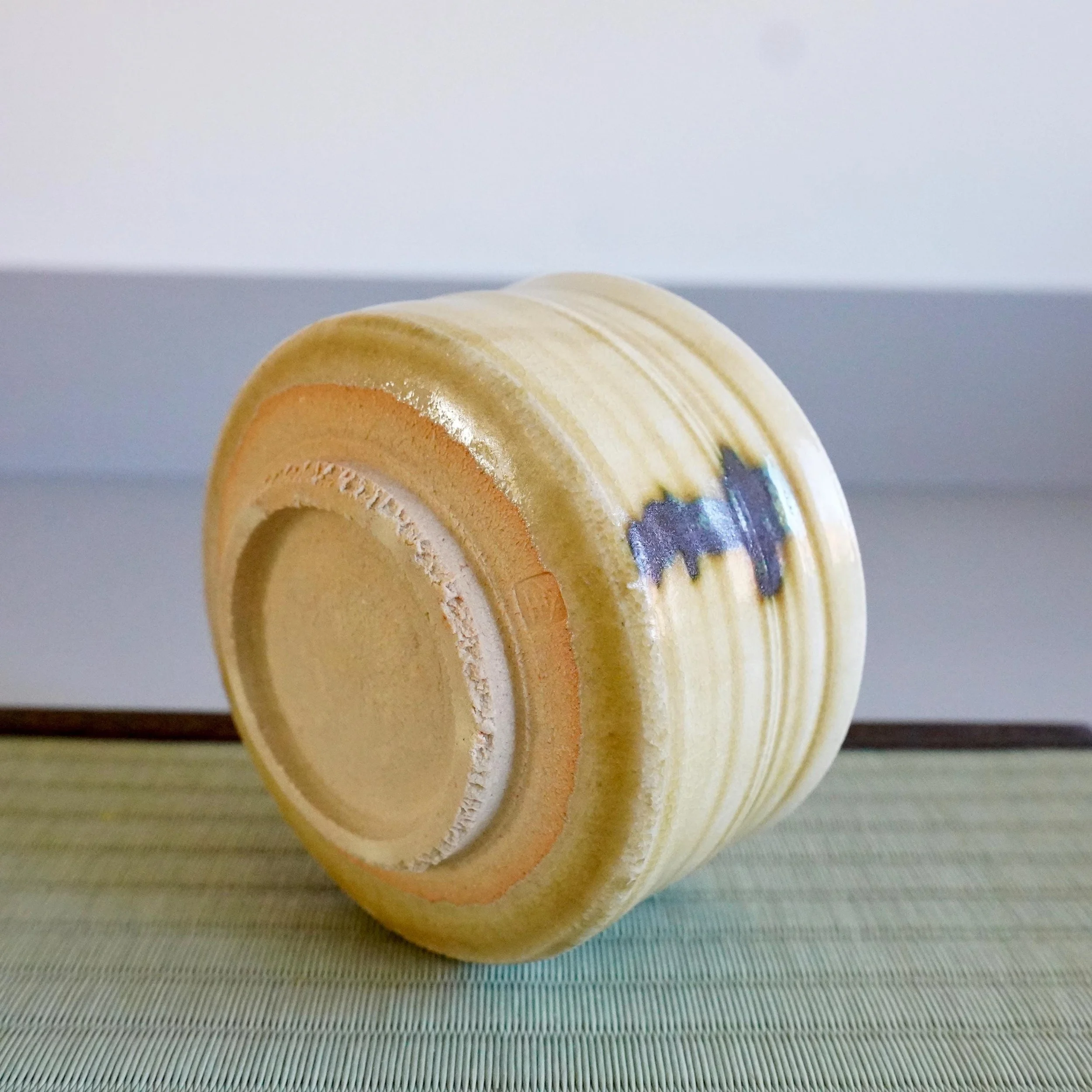 Yasuda Masahiro's Ki-Seto chawan, tilted on a tatami mat, displaying its ribbed mustard-yellow body with dark glaze drips and an impressed seal on the foot.