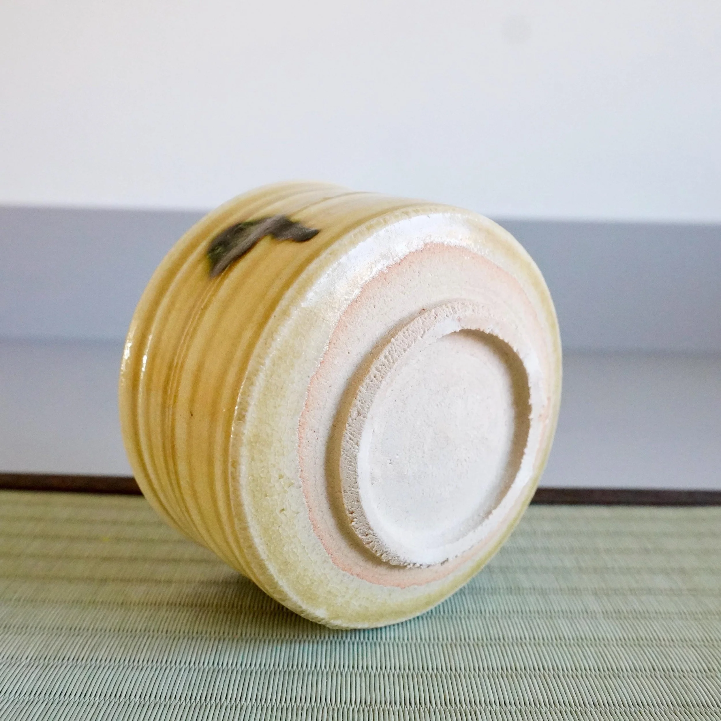 Ki-Seto chawan by Yasuda Masahiro, tilted to show its mustard-yellow ribbed side and unglazed foot with concentric rings, resting on a tatami mat.