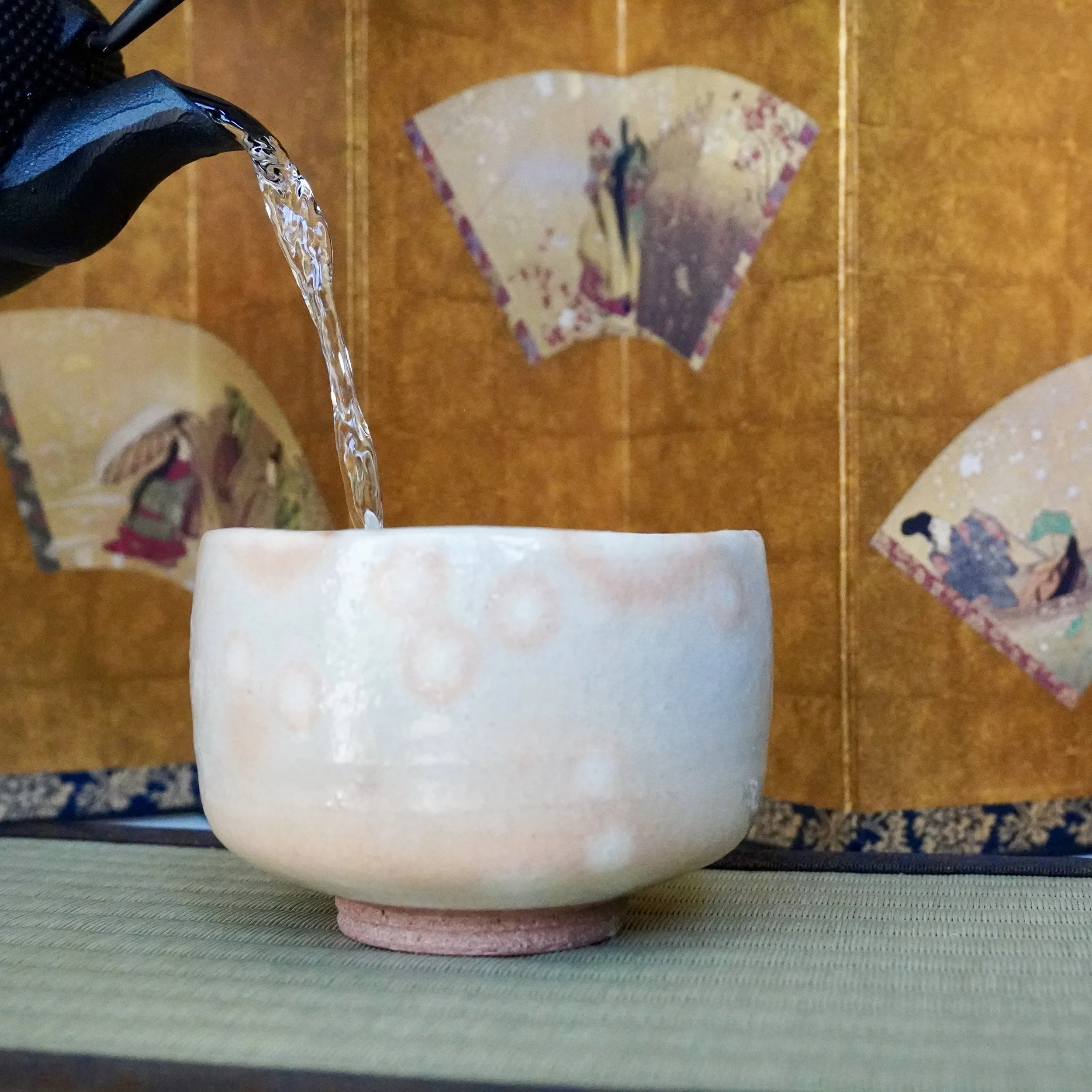 Water poured into Hagi-yaki chawan showing kan'nyū crackle glaze absorption, matcha bowl on tatami mat with gold folding screen background, Japan