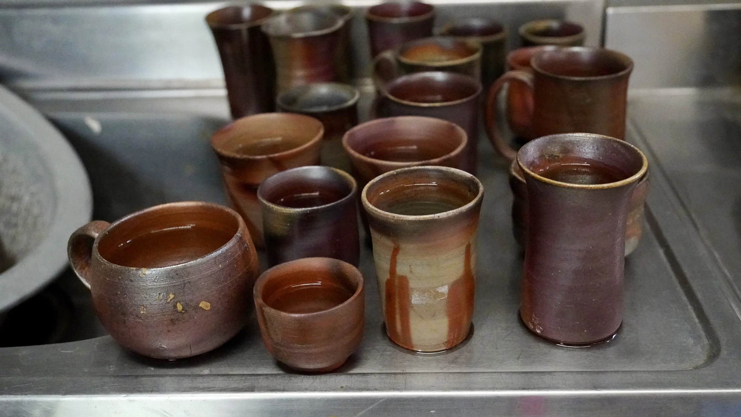 Freshly fired Bizen-yaki cups and mugs in warm red-brown and grey tones cooling on a workbench, Imbe Okayama Japan