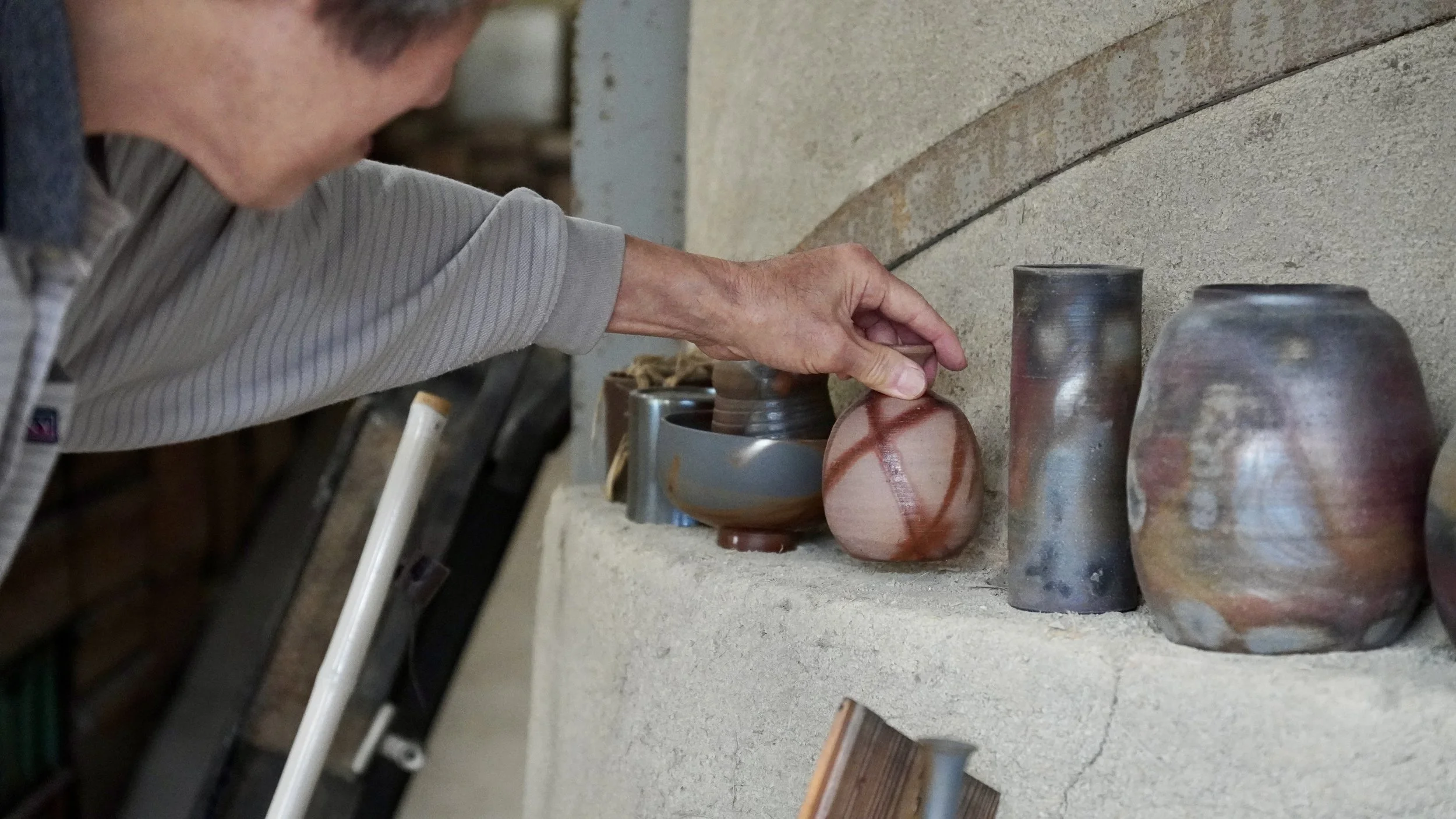 Hand of Shibaoka Masashi, third-generation Kōzan, reaching for a Bizen-yaki piece with hidasuki scarlet flame marks at Kōzan-gama kiln, Imbe Okayama Japan