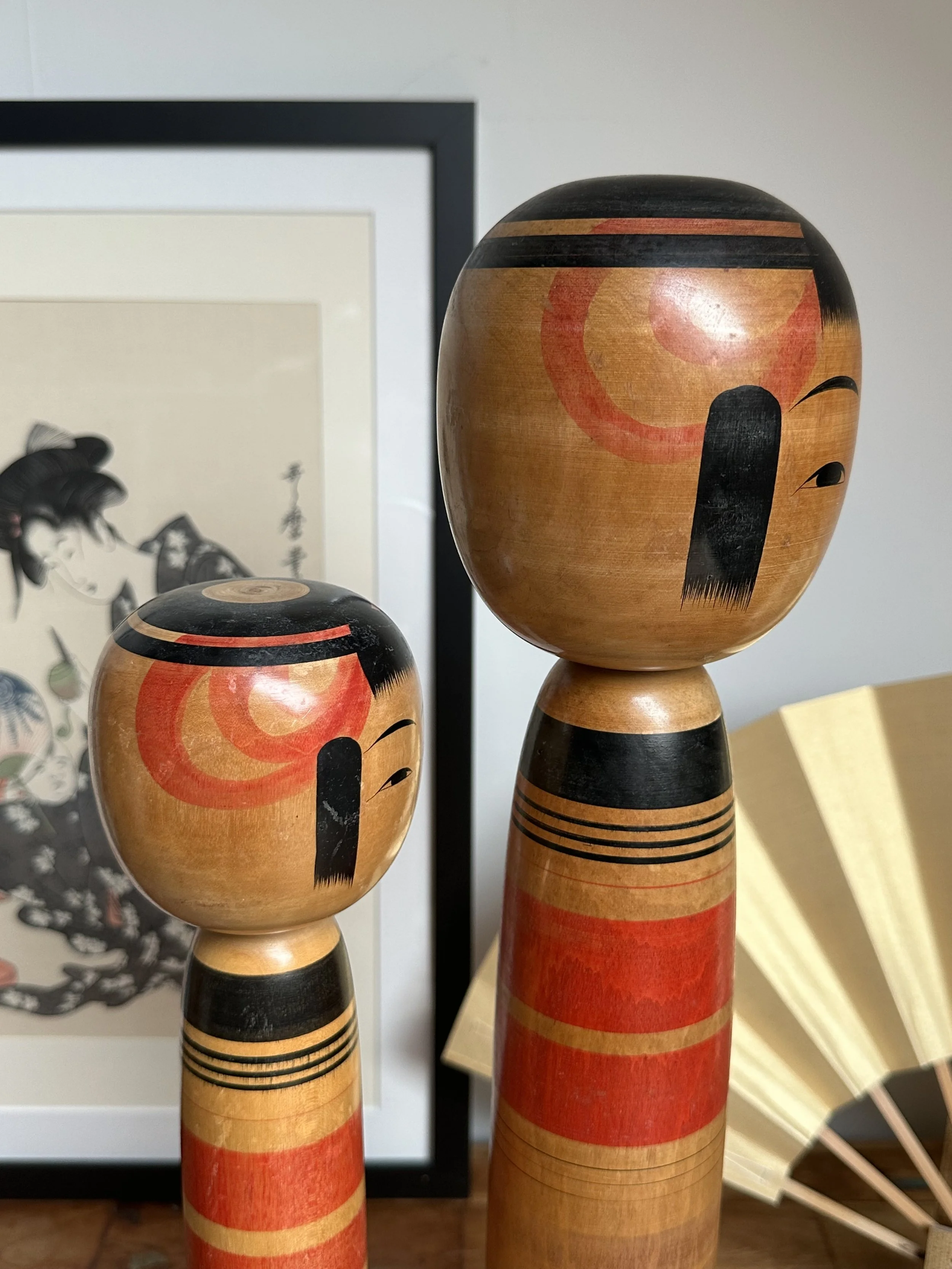 Two Tsuchiyu kokeshi by Abe Ichirō stand side-by-side, a taller and shorter doll. Both feature wooden bodies with red and black bands, and serene faces with black fringes and red circular marks.