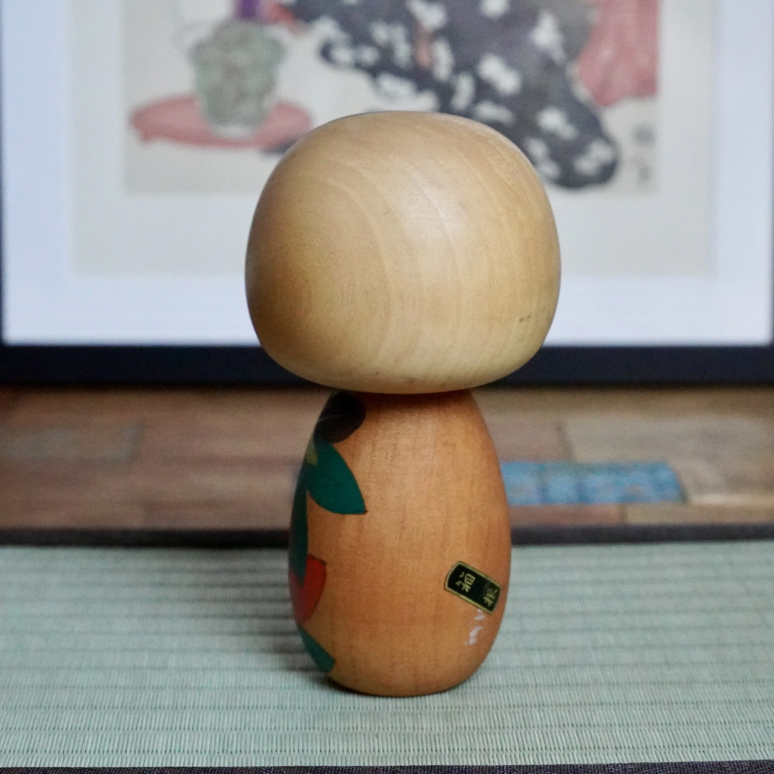 Back view of a kokeshi by Yuji Kawase, with a plain wooden head and a body carved and painted with green and red leaves. A black and yellow signature sticker is on its lower back.