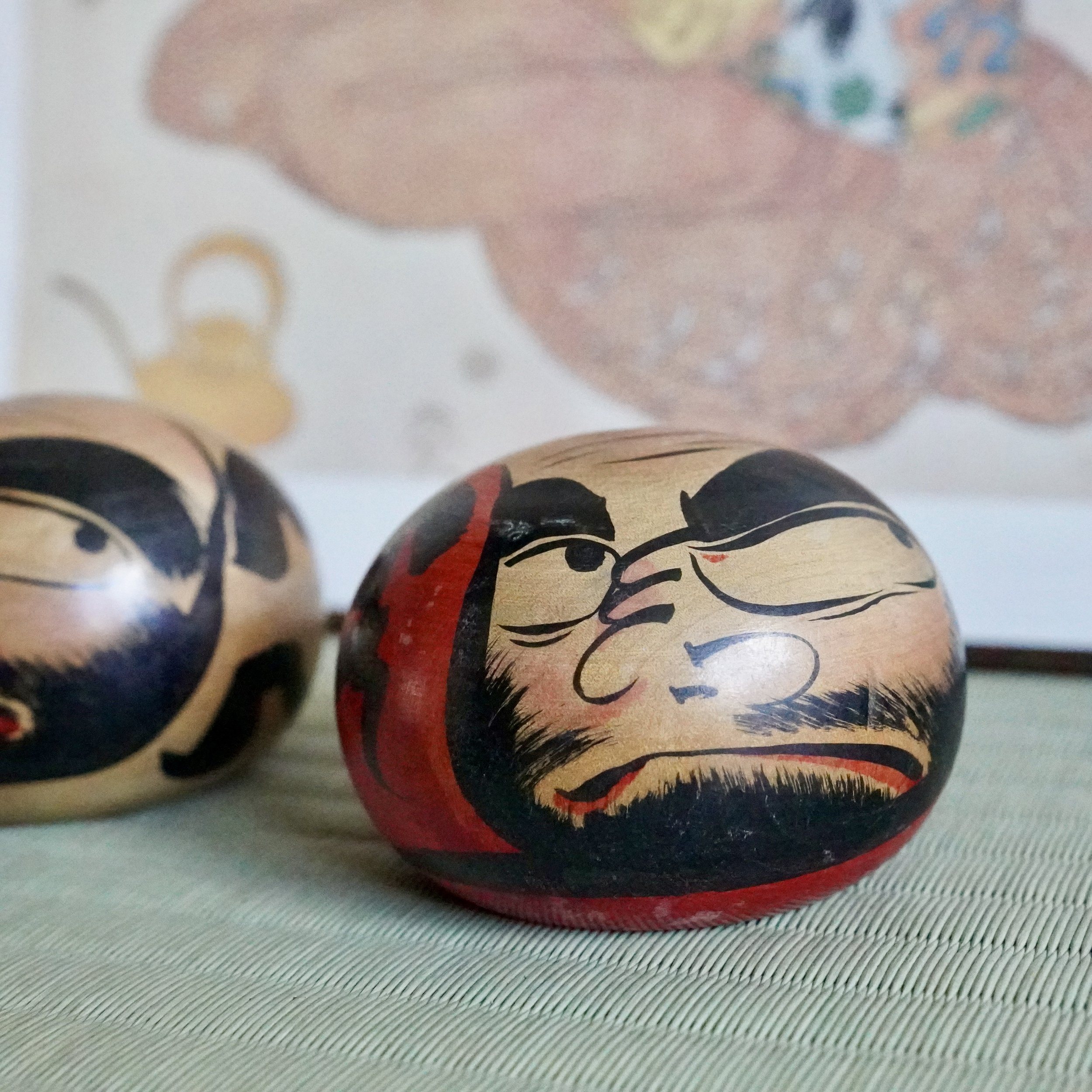 Hand-painted wooden Tsugaru daruma by Satō Zenji, Nuruyu. One doll in focus with scowling face, black beard, red collar, and a blurred second doll visible left.
