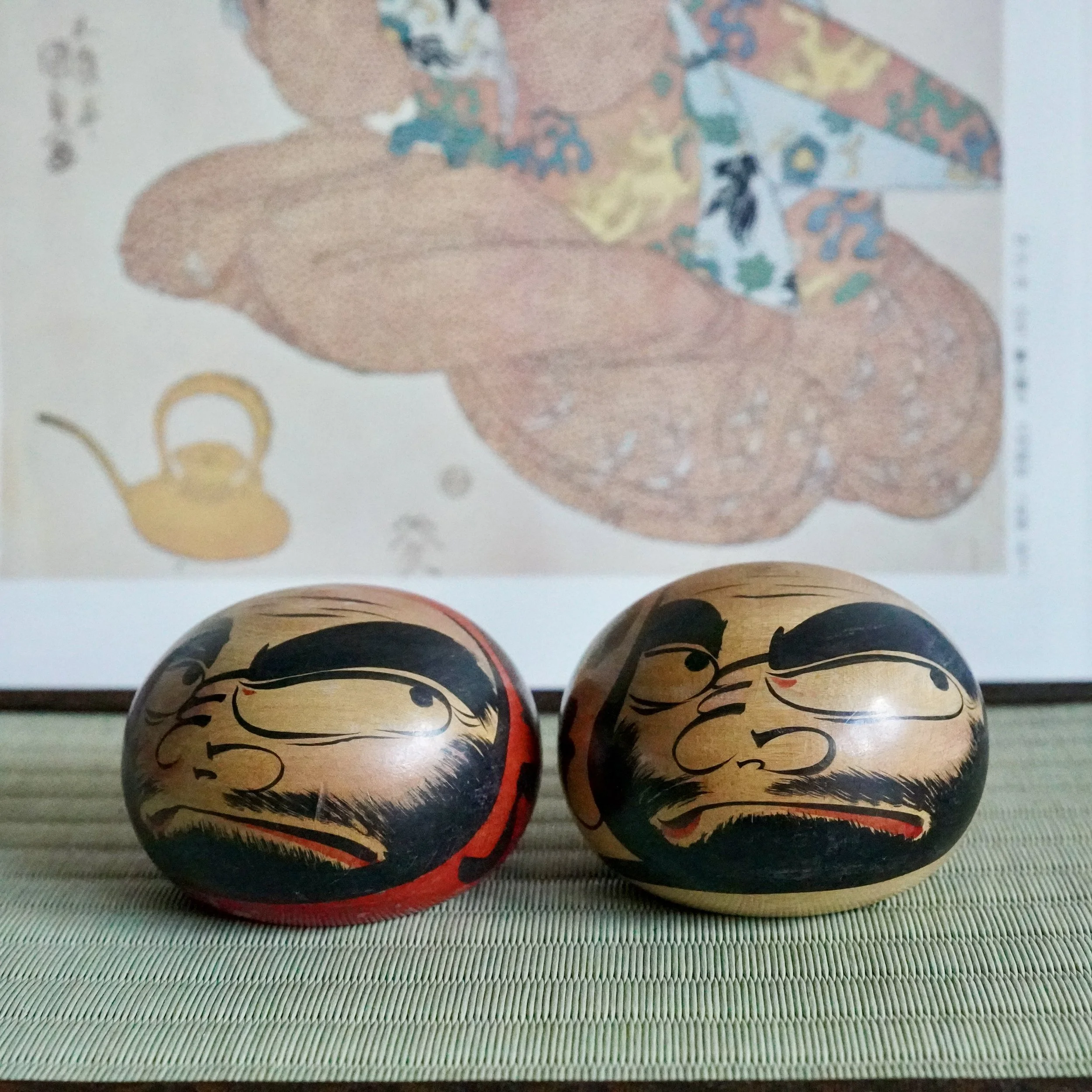 Pair of hand-painted wooden Tsugaru daruma caught mid-scowl, one with a red base, on a light green woven mat. Blurred background print.