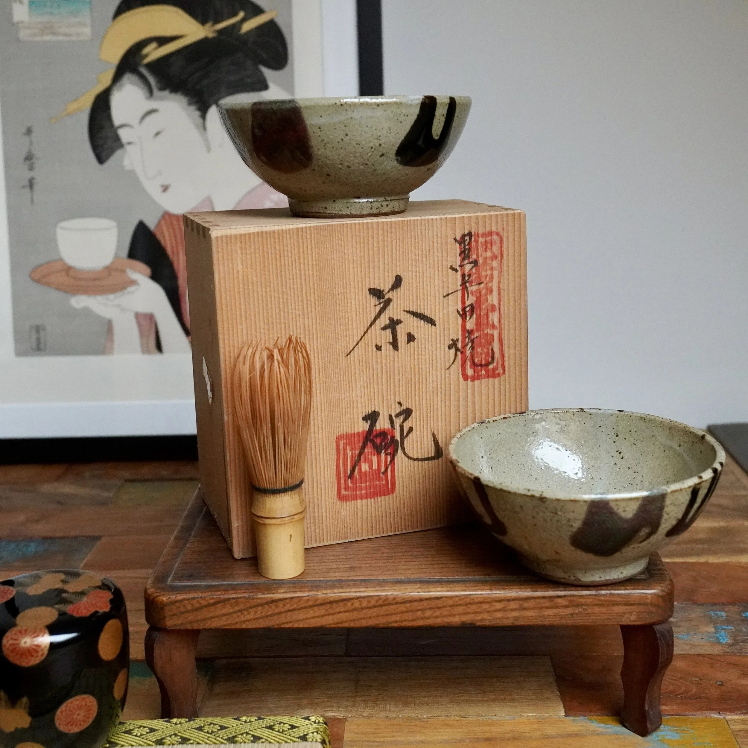 Two shallow, speckled tea bowls with dripping white, black, and reddish-brown glazes. One rests on an inscribed wooden box, the other by a bamboo whisk, with an ukiyo-e print behind.