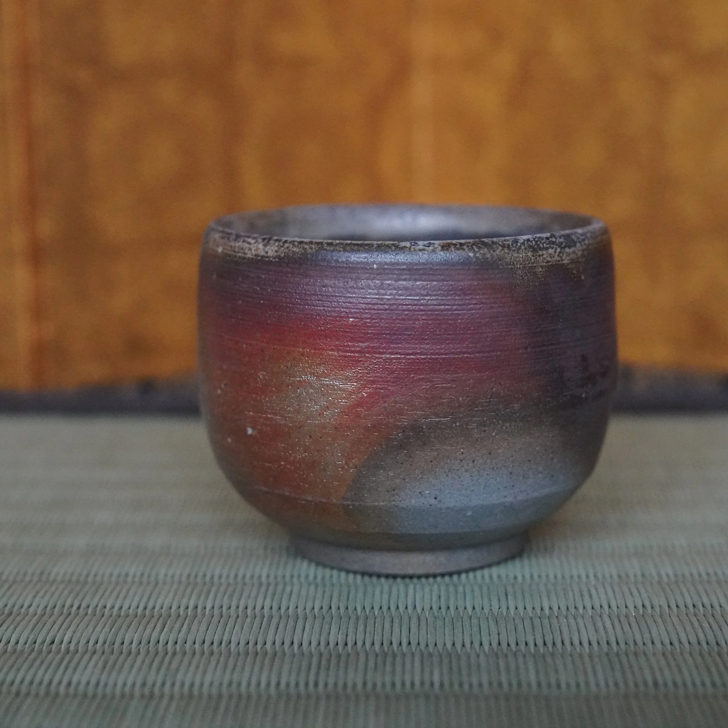 Yamamoto Sōshū Bizen-yaki guinomi, hand-thrown with dark reddish-brown and scarlet hidasuki flame marks, on a woven mat.