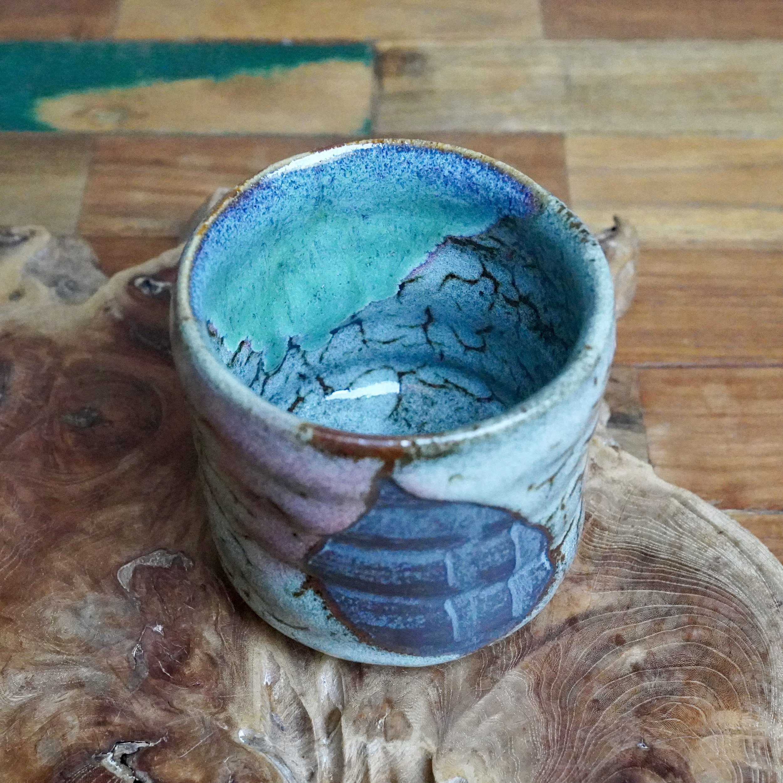 Tall tsutsu chawan with a vibrant turquoise interior transitioning to a cracked blue-grey. Its exterior features similar glaze with brown lines and subtle pink patches, on wood.