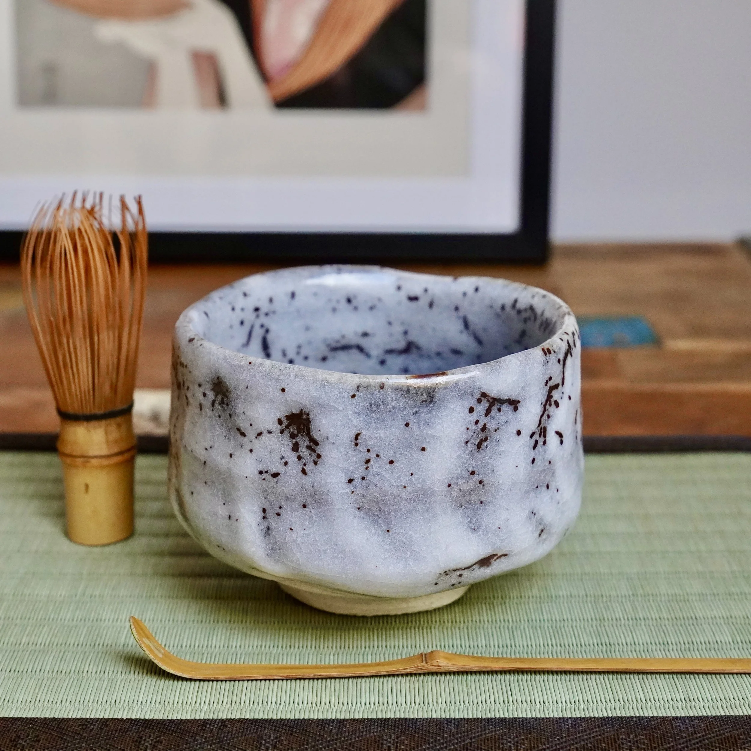 Katō Shōzan's Oni-Shino chawan, rustic white with rust brown speckles and textured walls, alongside a bamboo whisk and scoop on a green tatami mat.