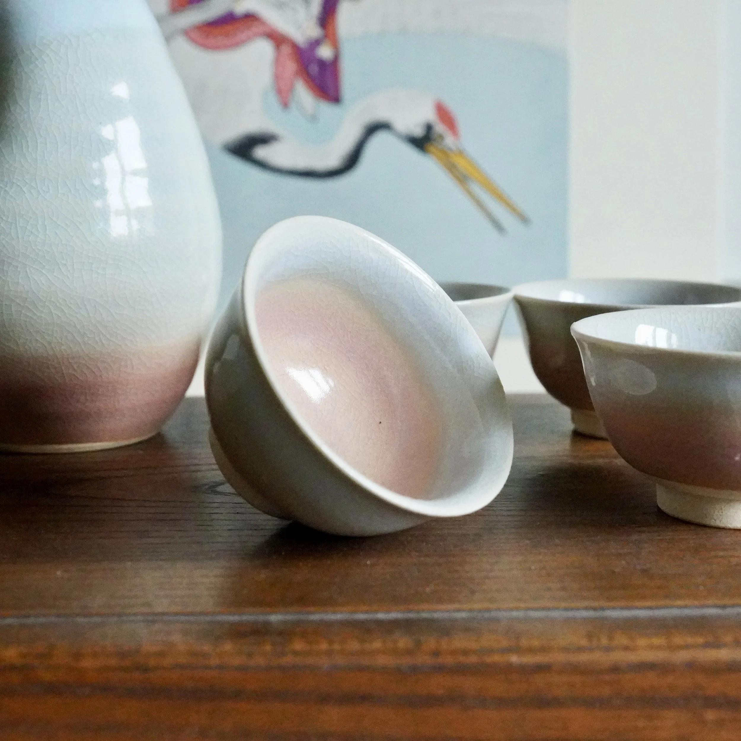 Hagi-yaki sake set: tokkuri and ochoko on wood. Gradient crackle glaze from blush pink to pale blue-grey. Foreground cup tilted.
