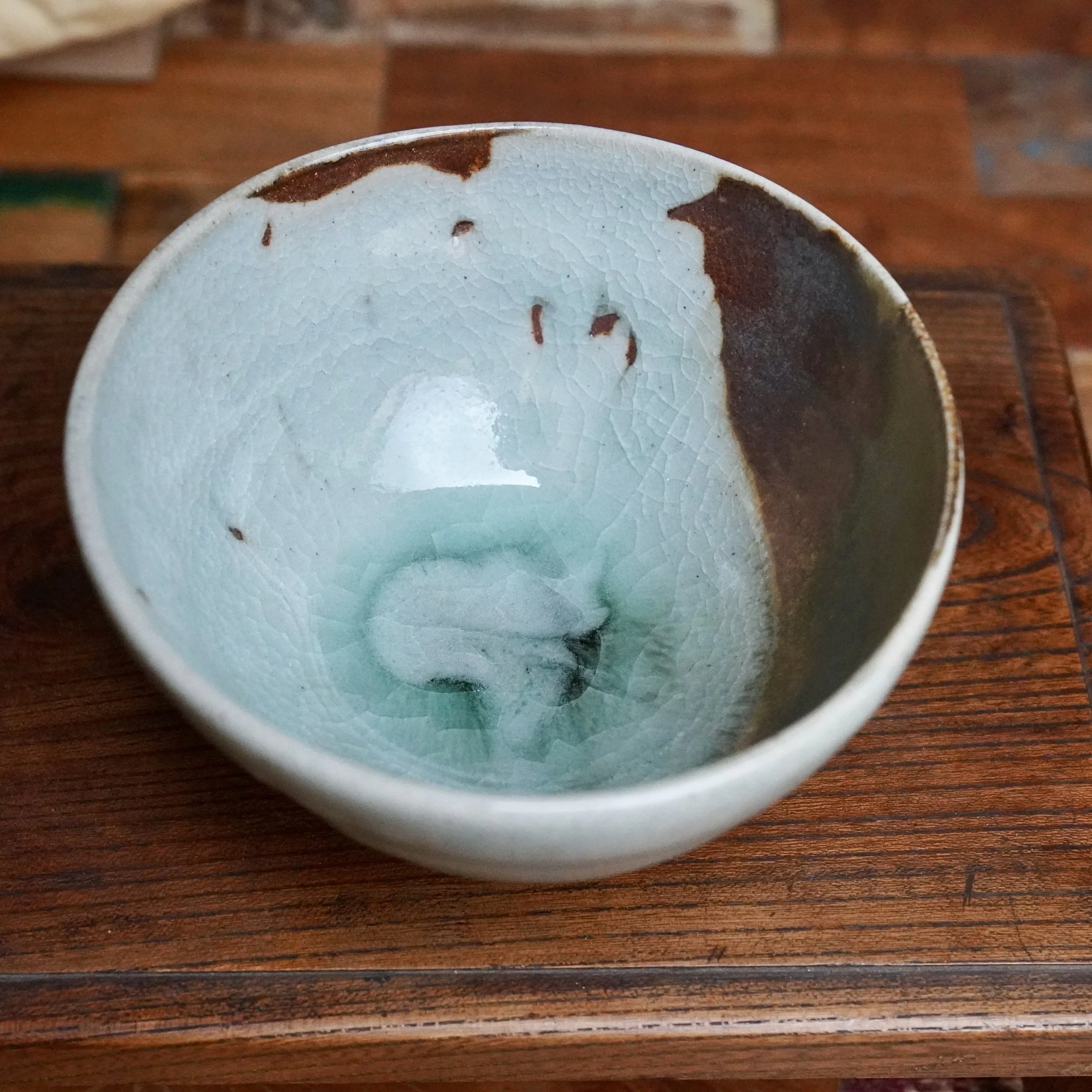 Chawan with a light blue-green ice crackle glaze inside, flowing into a rich rust-brown on one side, on a dark wooden surface.
