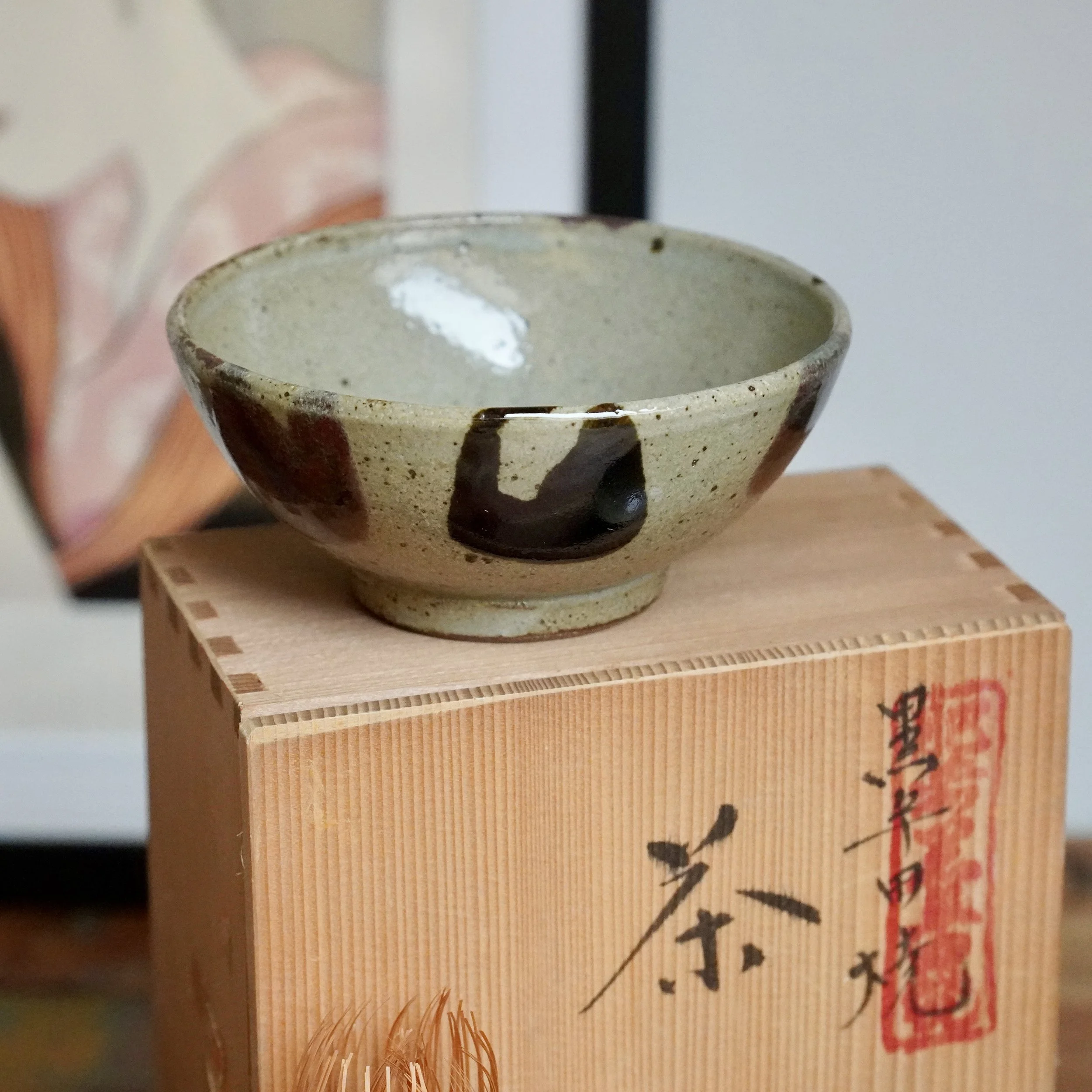 Shallow tea bowl by Maruta Nobumasa kiln with a speckled pale glaze, dark brush strokes, and reddish tones, resting on a wooden tomobako with black calligraphy and a red seal.