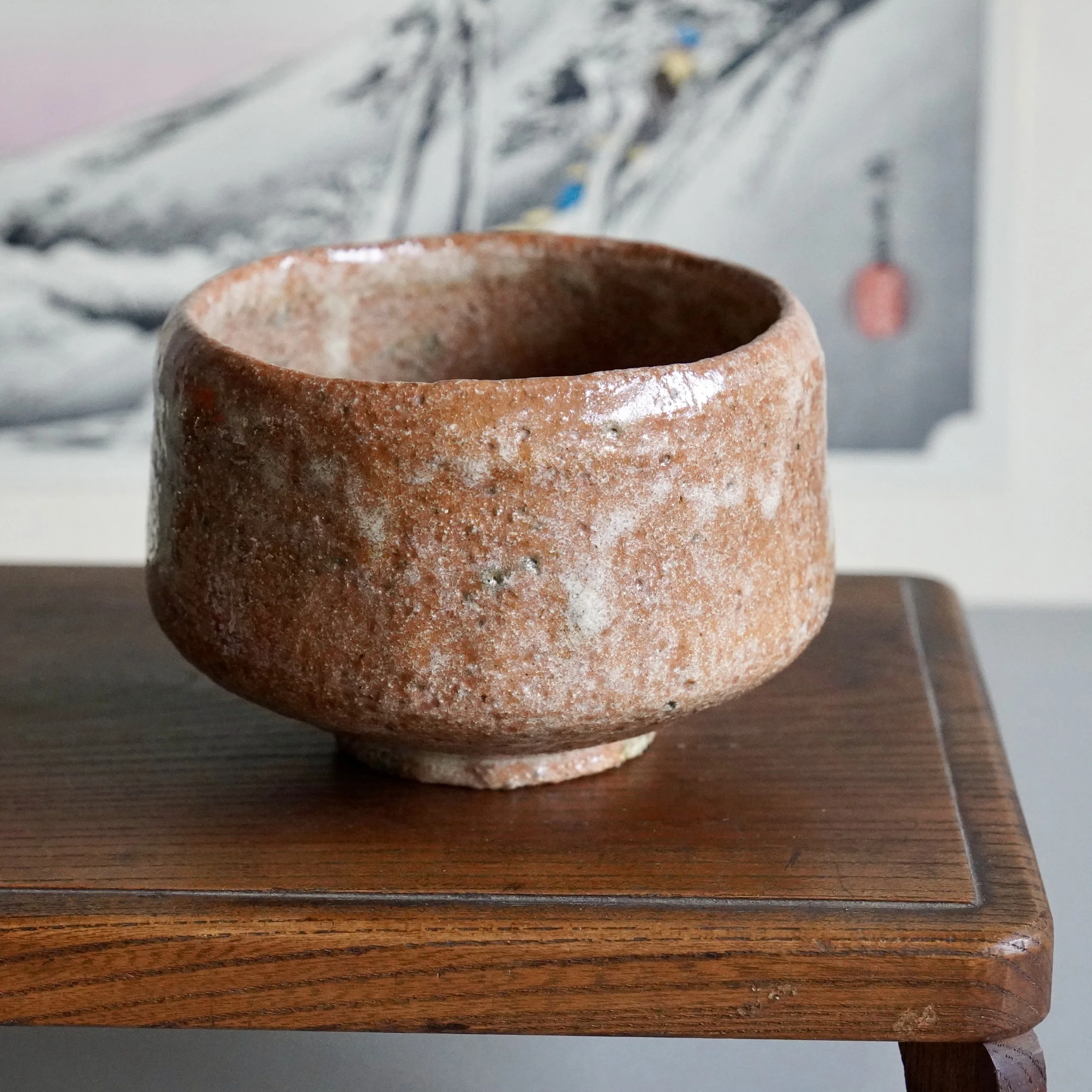 Sawada Hiroyuki aka-raku chawan, rugged reddish-brown, with sandy, pitted texture and a prominent white glaze drip, on a dark wooden stand.