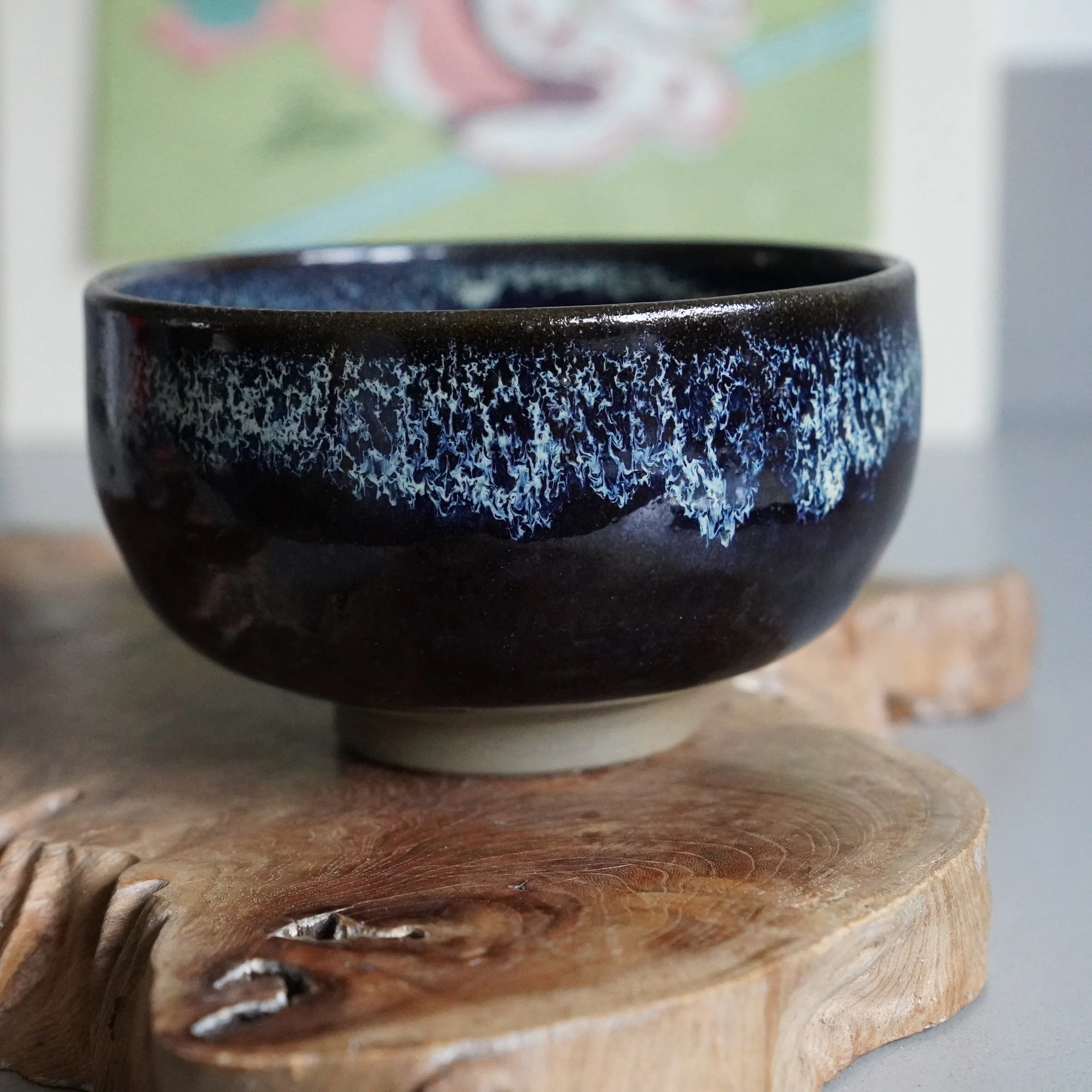 Ceramic matcha bowl with dark brown glaze and mottled blue-white patterns on the rim, resting on a wooden slice.