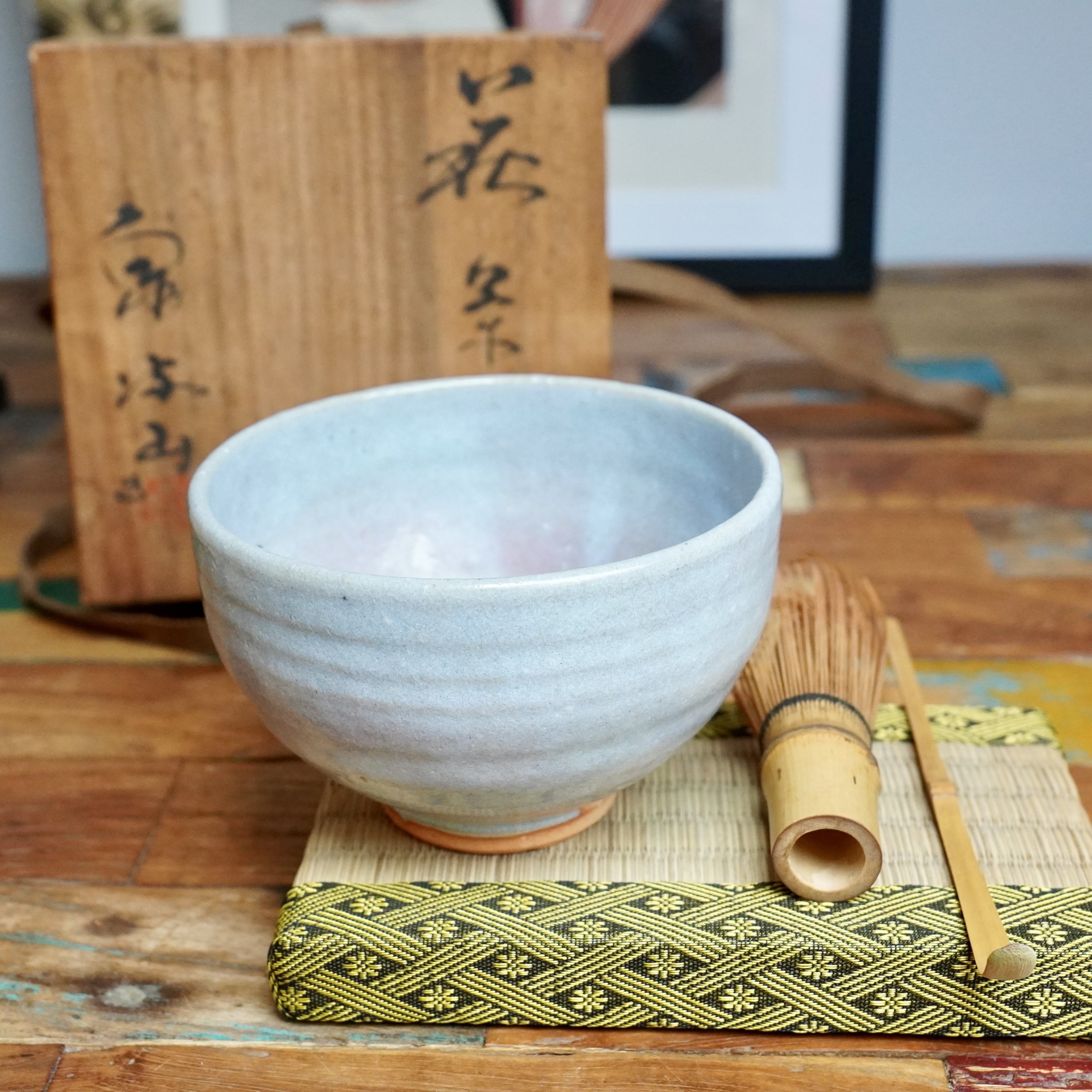 Hagi-yaki chawan by Moriwaki Fumitada: a low, pale blue-grey ceramic bowl, with a bamboo whisk, scoop, and patterned mat, and a calligraphed wooden box behind.