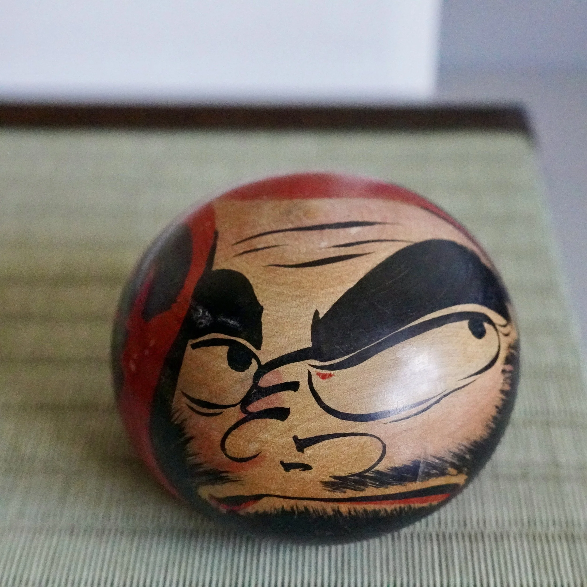 Tsugaru daruma by Satō Zenji, a hand-painted wooden doll with a scowling face, thick black eyebrows, mustache, and beard, accented with red, on a woven mat.