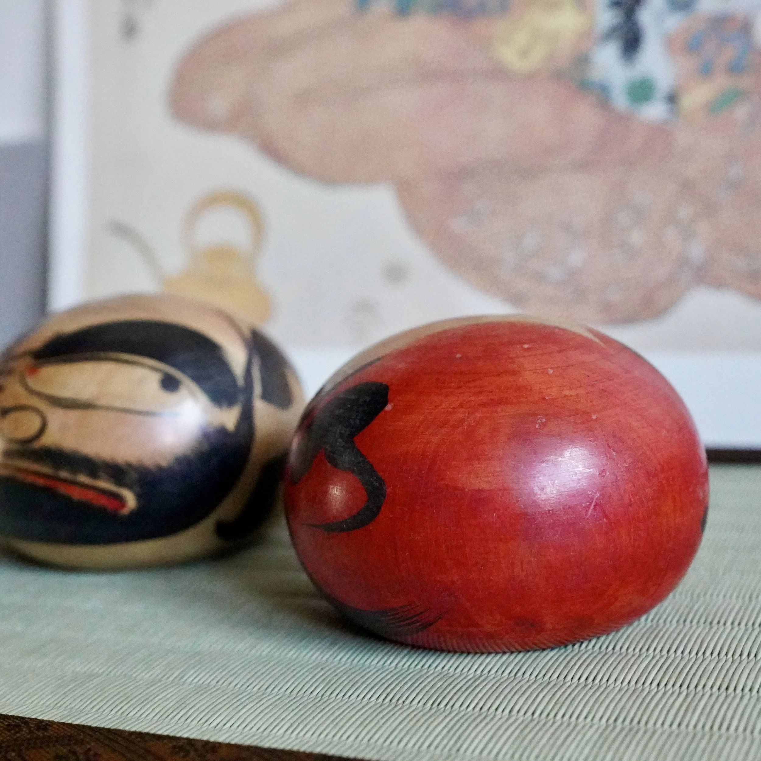 Two round daruma dolls on a textured light green mat. The foreground doll is red with a black swirl; the other is natural wood with a painted scowling face.
