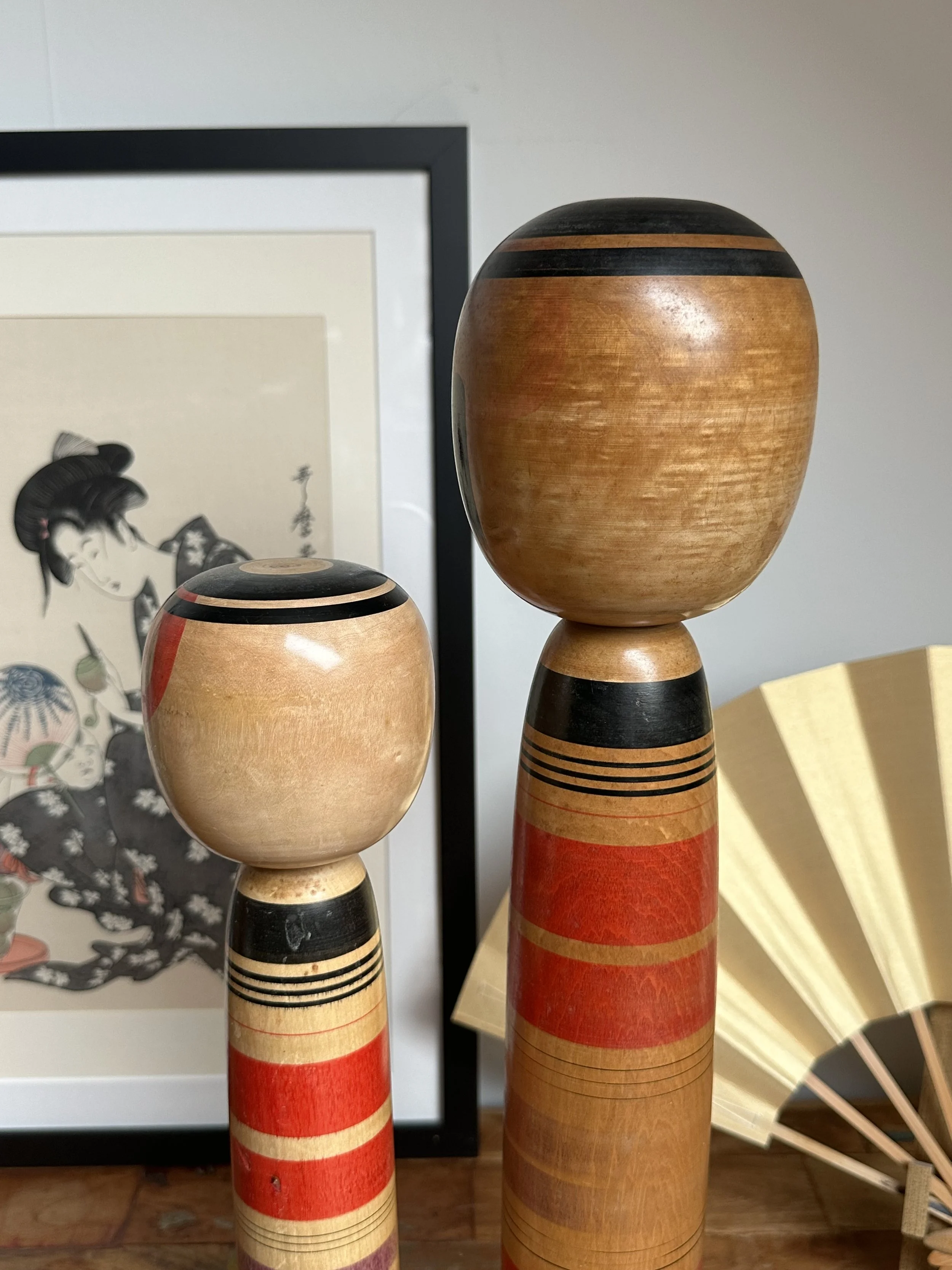 Two Abe Ichirō Kokeshi dolls, one taller, with smooth wood bodies and red, black, and natural horizontal bands. Framed print and fan in the background.