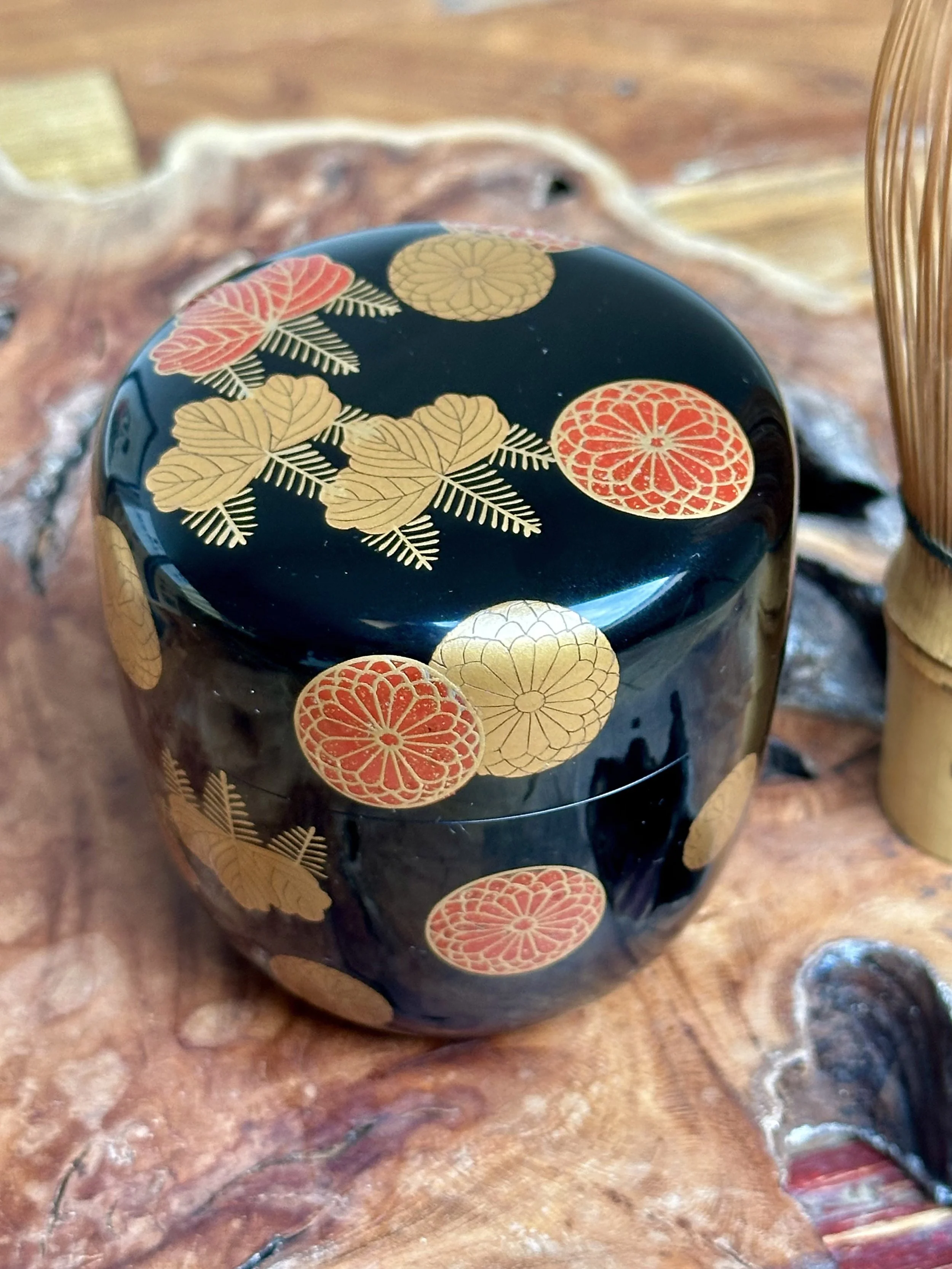 Glossy black natsume tea caddy with matte gold paulownia and flecked red chrysanthemum crests, on a dark wood surface with a blurred bamboo whisk.