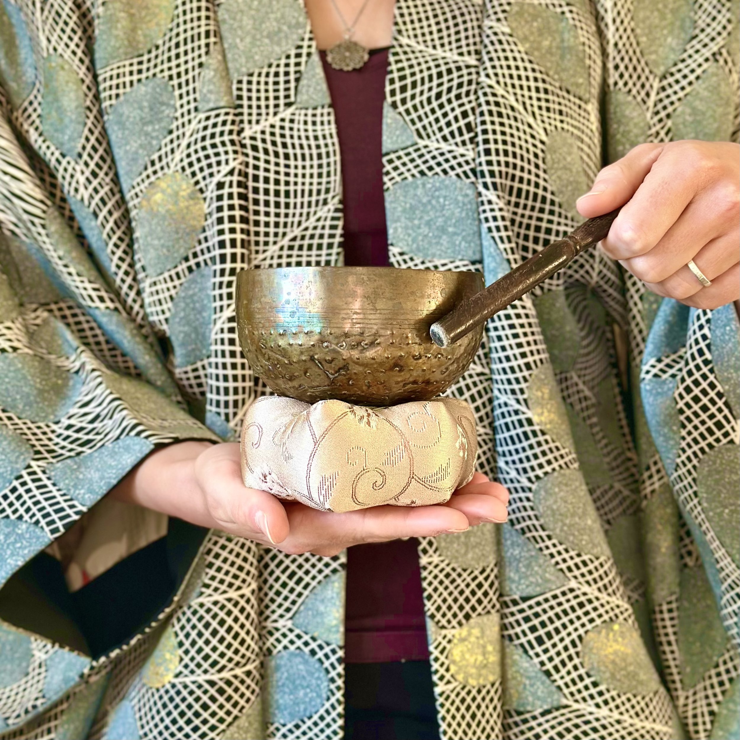 Close-up of a person wearing a patterned silk vintage haori kimono jacket and holding an antique brass singing bowl with a wooden mallet.