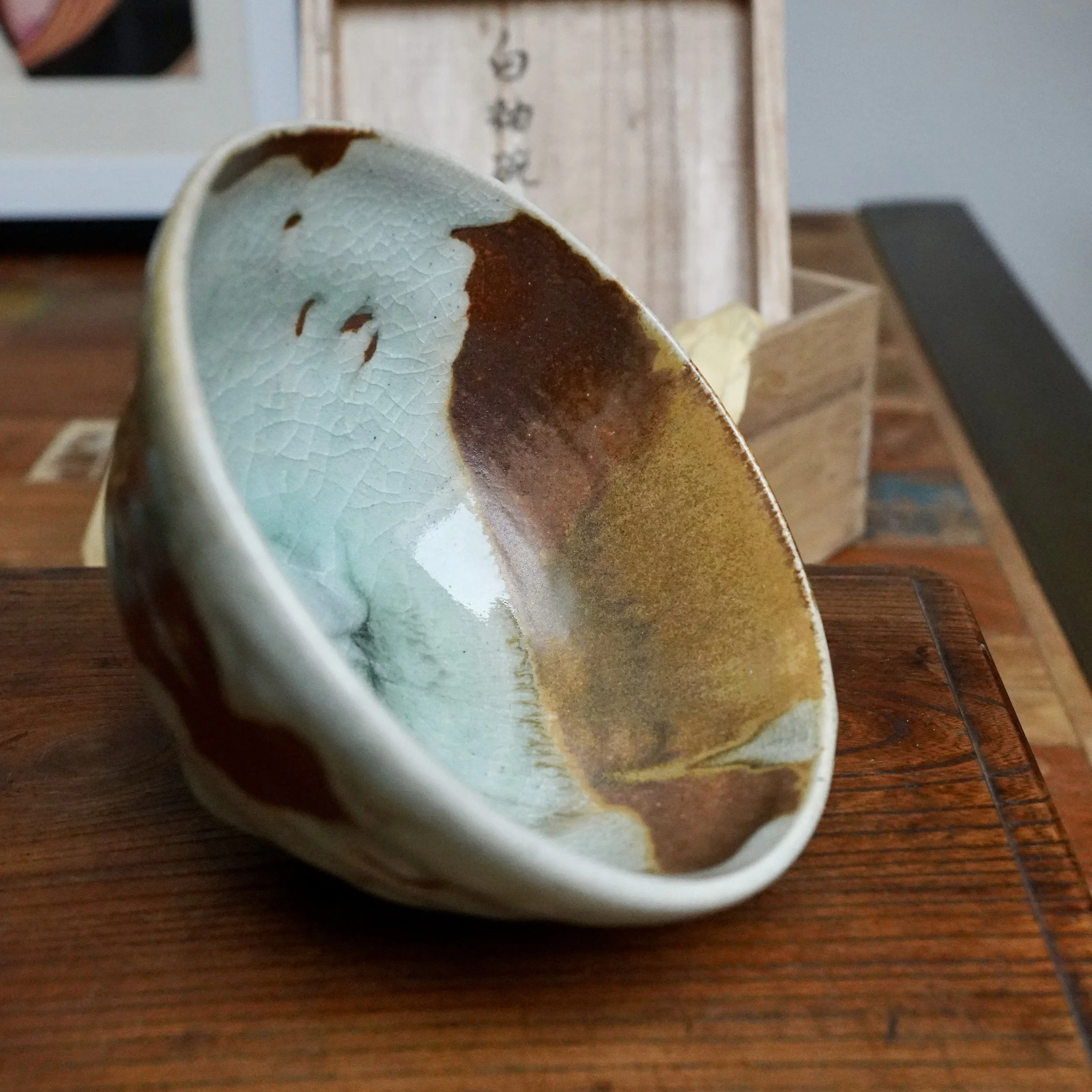 Chawan tilted on a wooden surface, showing its interior where pale, crackled blue-grey glaze transitions into deep rust-brown with ochre streaks. Blurred wooden box, signed Sansōan.