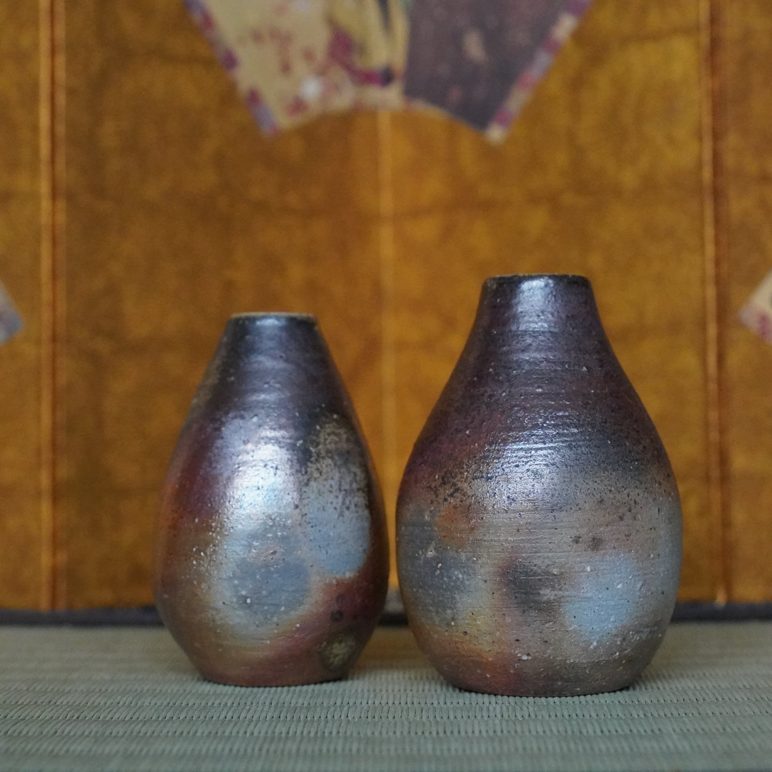 Shibaoka Tōsendō Bizen-yaki tokkuri pair, dark textured vases with silvery-blue and reddish-brown firing marks, on a woven mat.