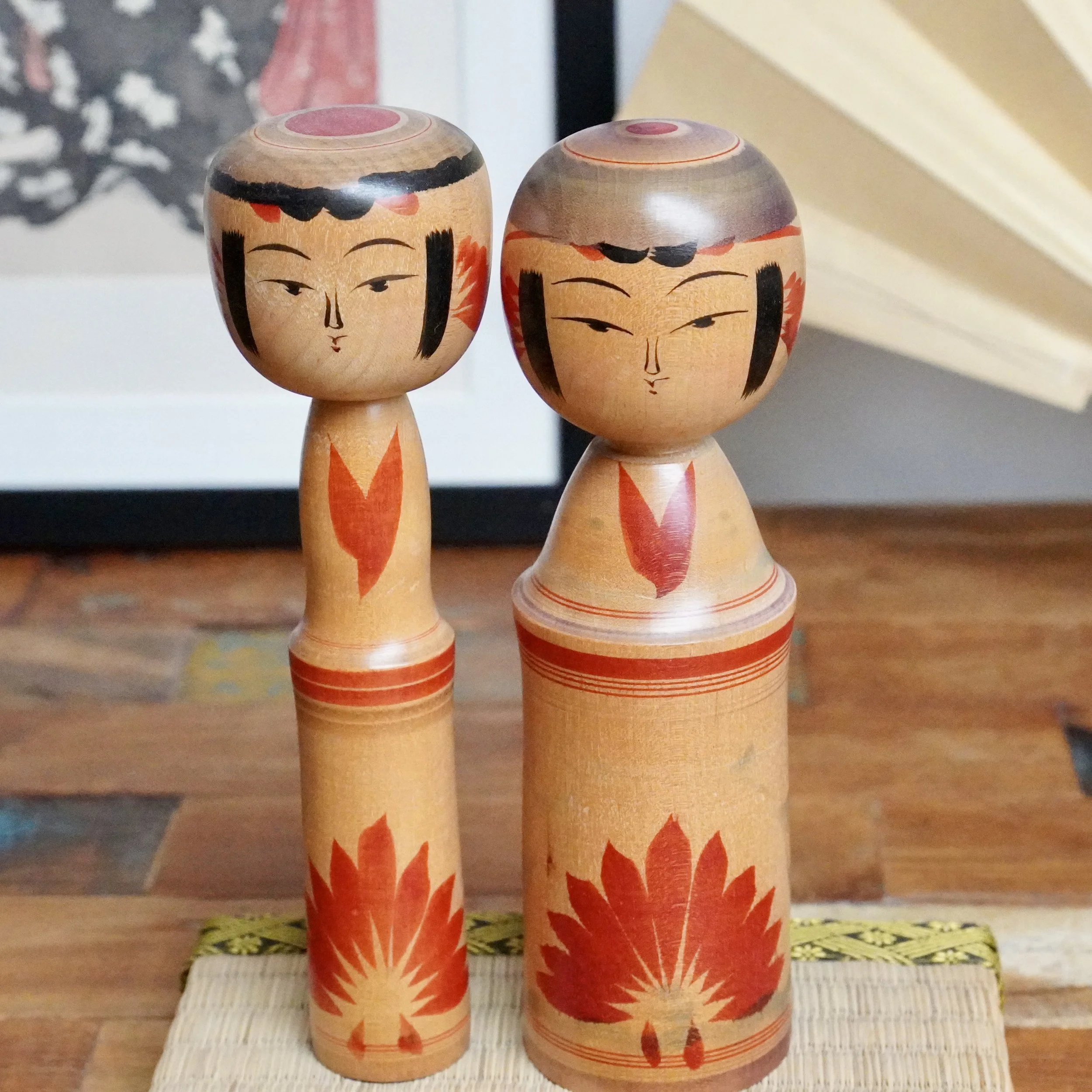 Pair of wooden kokeshi dolls by Inoue Harumi, standing side-by-side with painted faces, black hair, and red chrysanthemum motifs on their bodies, on a woven mat.