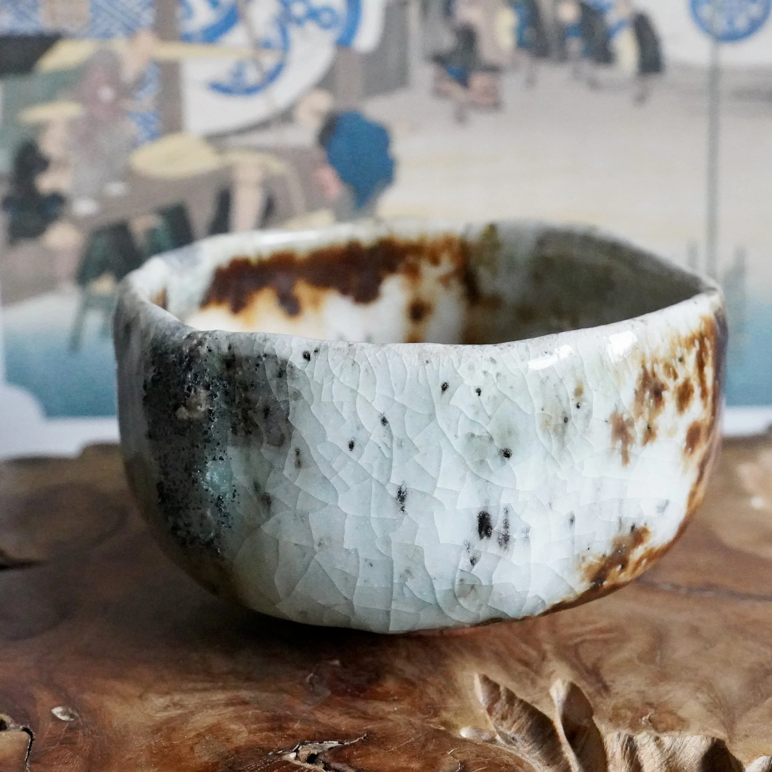 Deguchi Eiji chawan, its crackled pale blue-green glaze blending into rusty brown sections, on carved wood with blurred artwork behind.