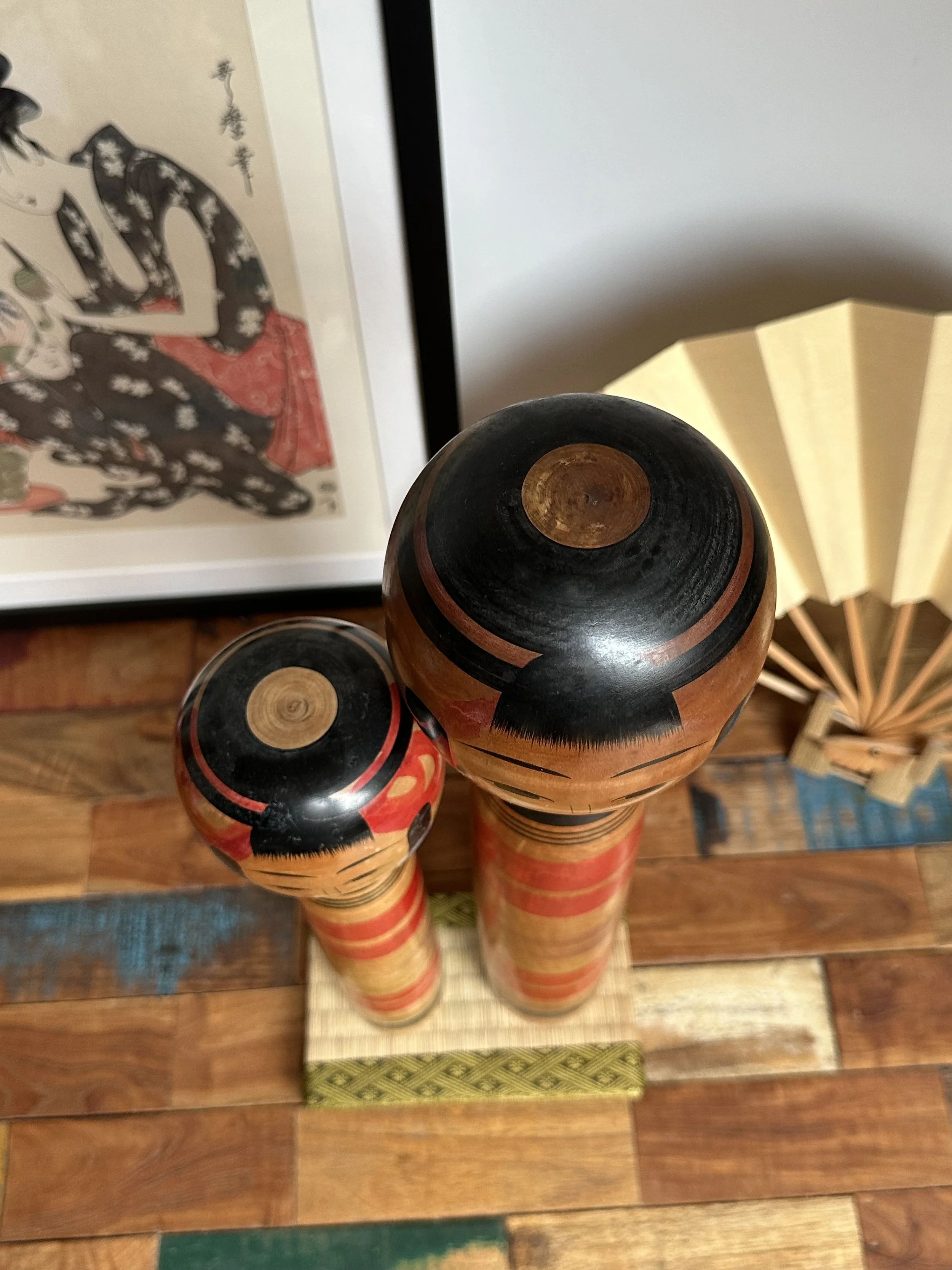 Top-down view of two Abe Ichirō Tsuchiyu kokeshi. Their black heads with concentric wood and red bands, and red-striped bodies, stand on a mat before a framed print and fan.