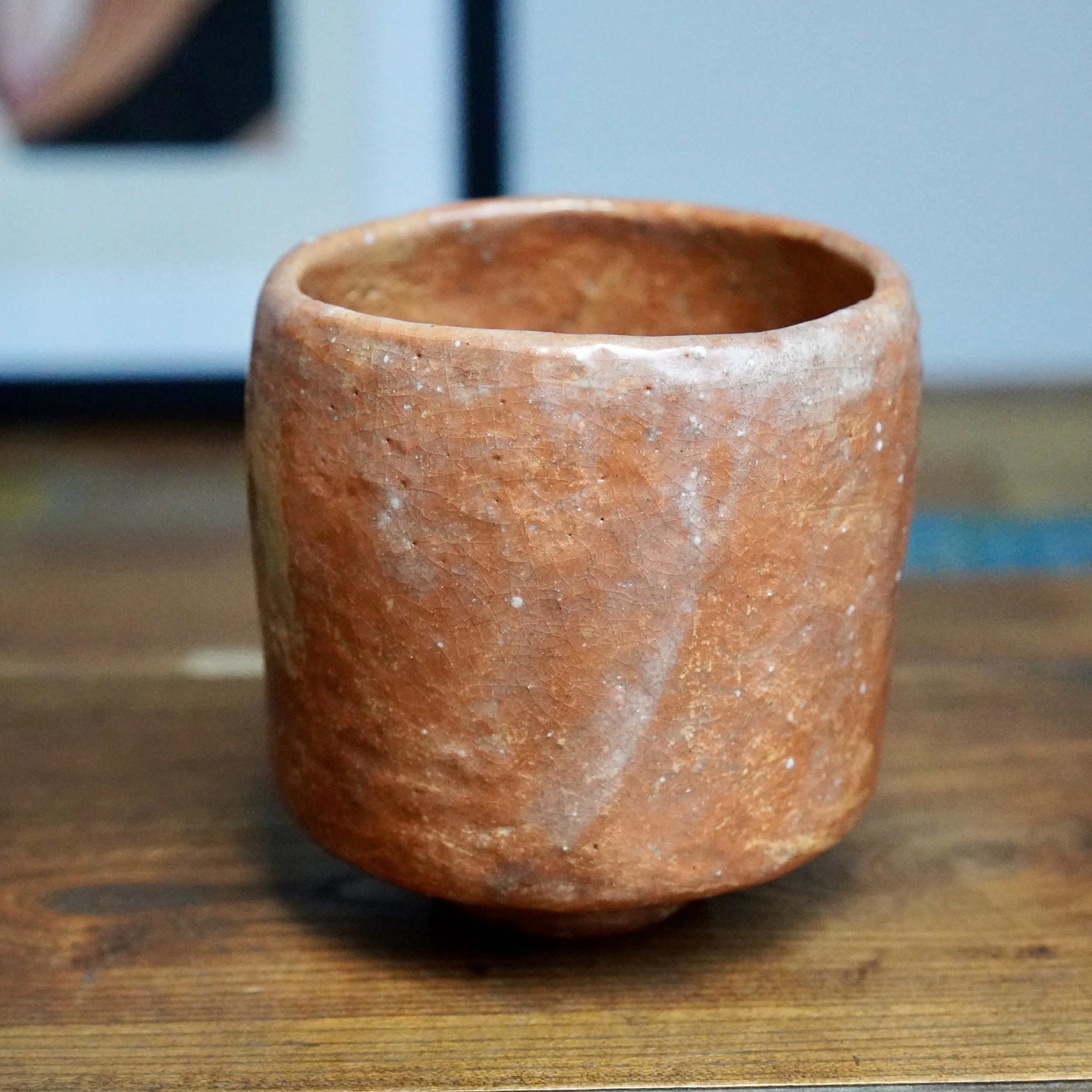Tall, terracotta-coloured tsutsu chawan with a crackle glaze and white drip marks from the rim, on a wooden surface.