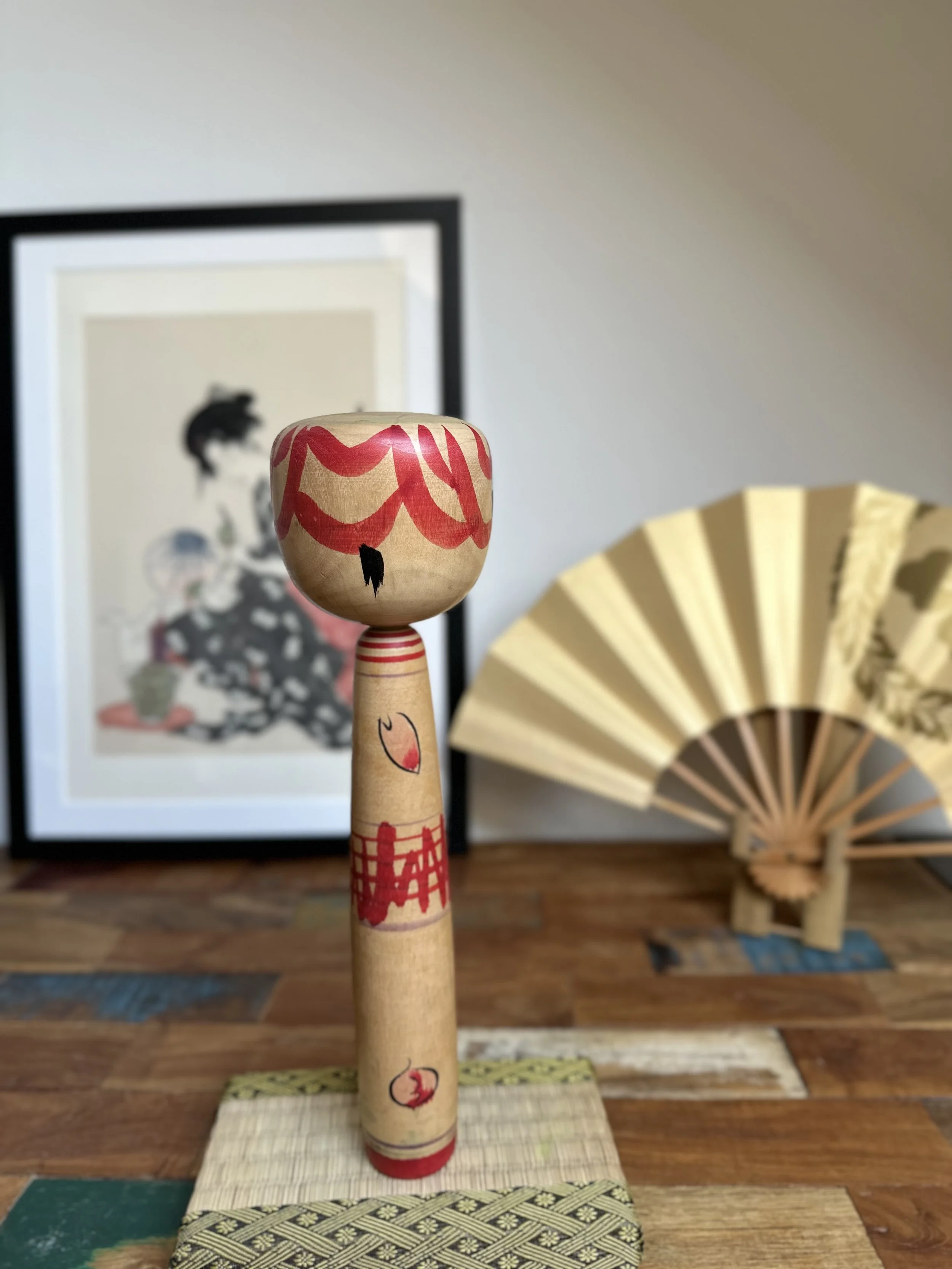 Seya Jūji's Nakanosawa Takobozu kokeshi doll, a wooden figure with a broad head, red wavy lines and a black mark, and a narrow body with red patterns, on a mat.