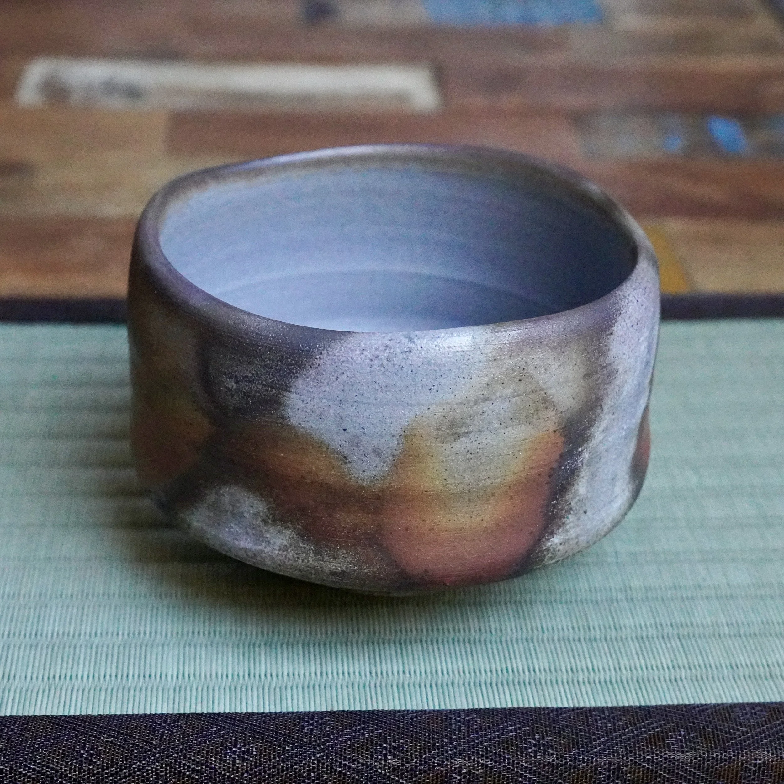 Inomata Masaaki Bizen chawan, textured with rust, orange, and ash grey firing effects, smooth blue-grey interior, on a green tatami mat.