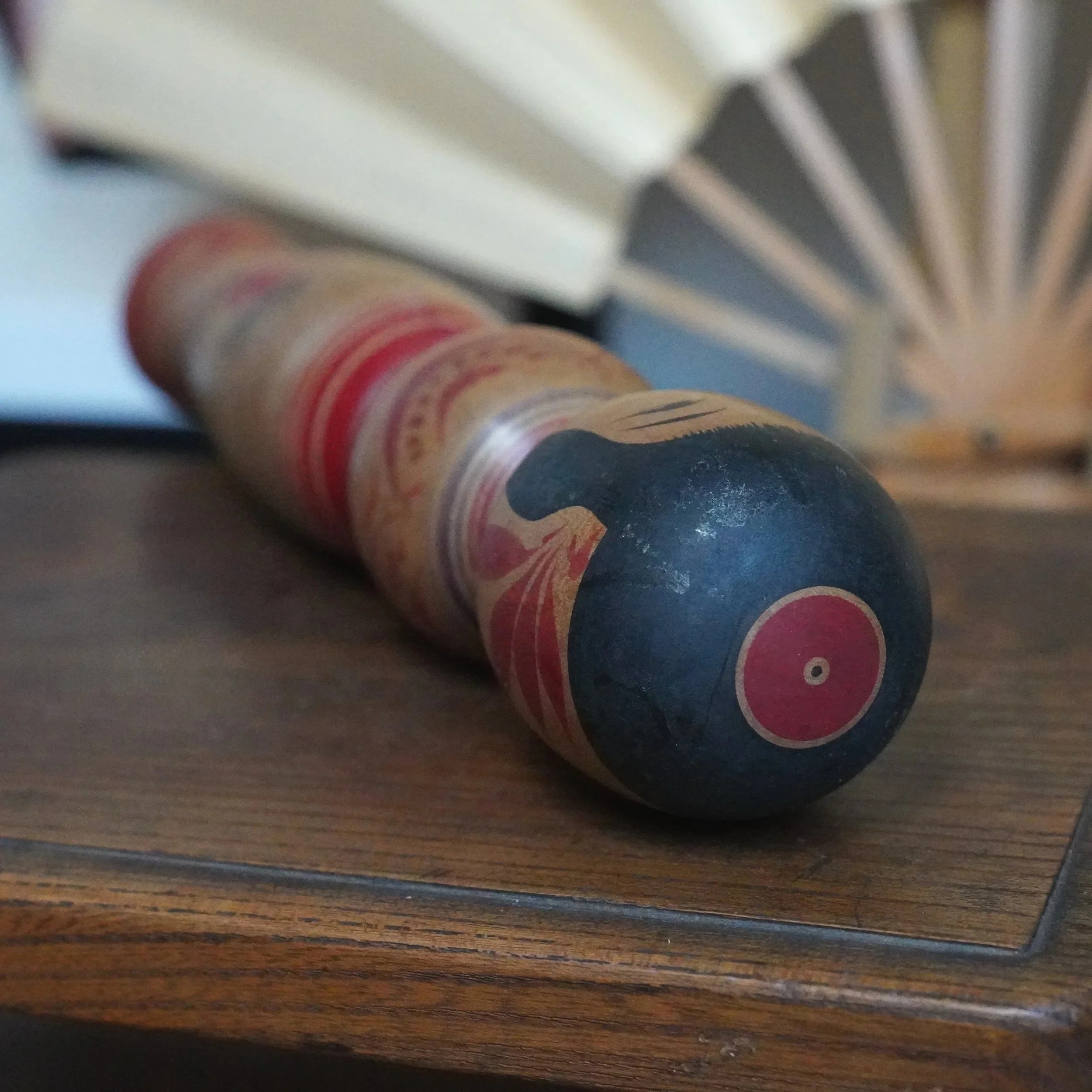 Tsugaru kokeshi doll by Sasamori Junichi, lying on a dark wood table. It features a dark round head with a red circle and central black dot, and a wood body with sweeping red peony designs.