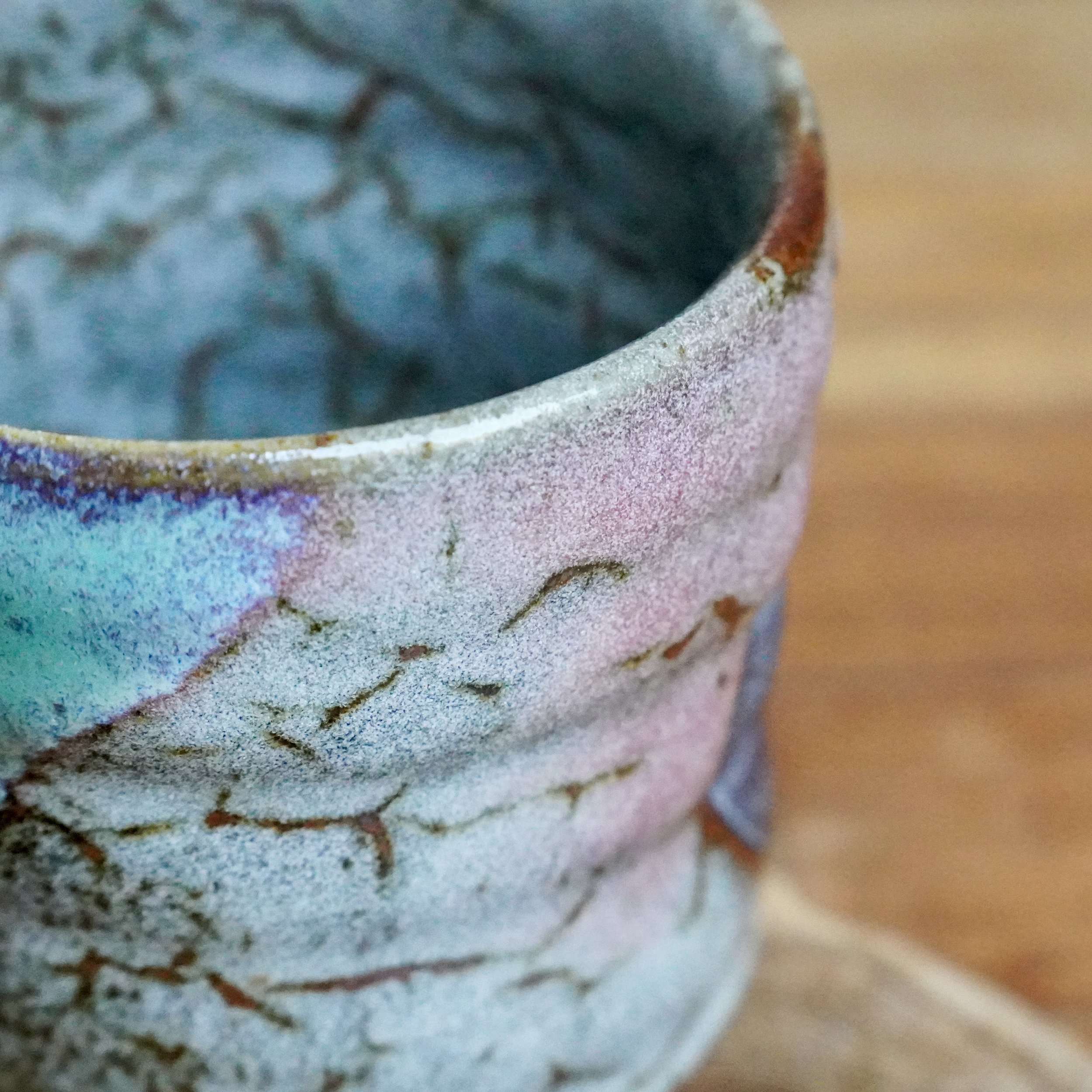 Close-up of a chawan with layered turquoise and speckled grey glaze, featuring warm brown lines and a blurred wooden background.