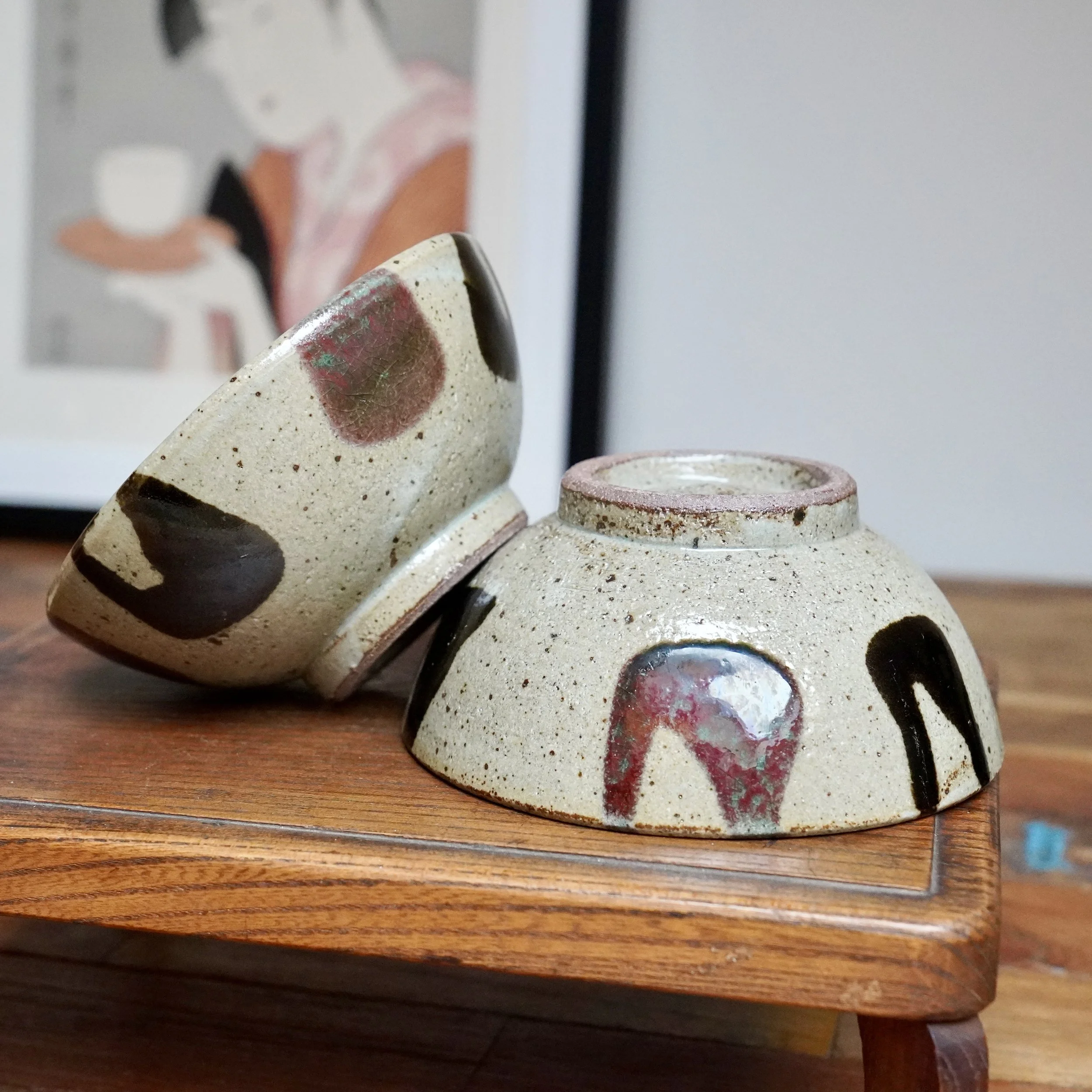 Two Maruta Nobumasa Kuromuta-yaki bowls, inverted on wood, showing speckled clay with white drips, black strokes, and wine-red glaze patches.
