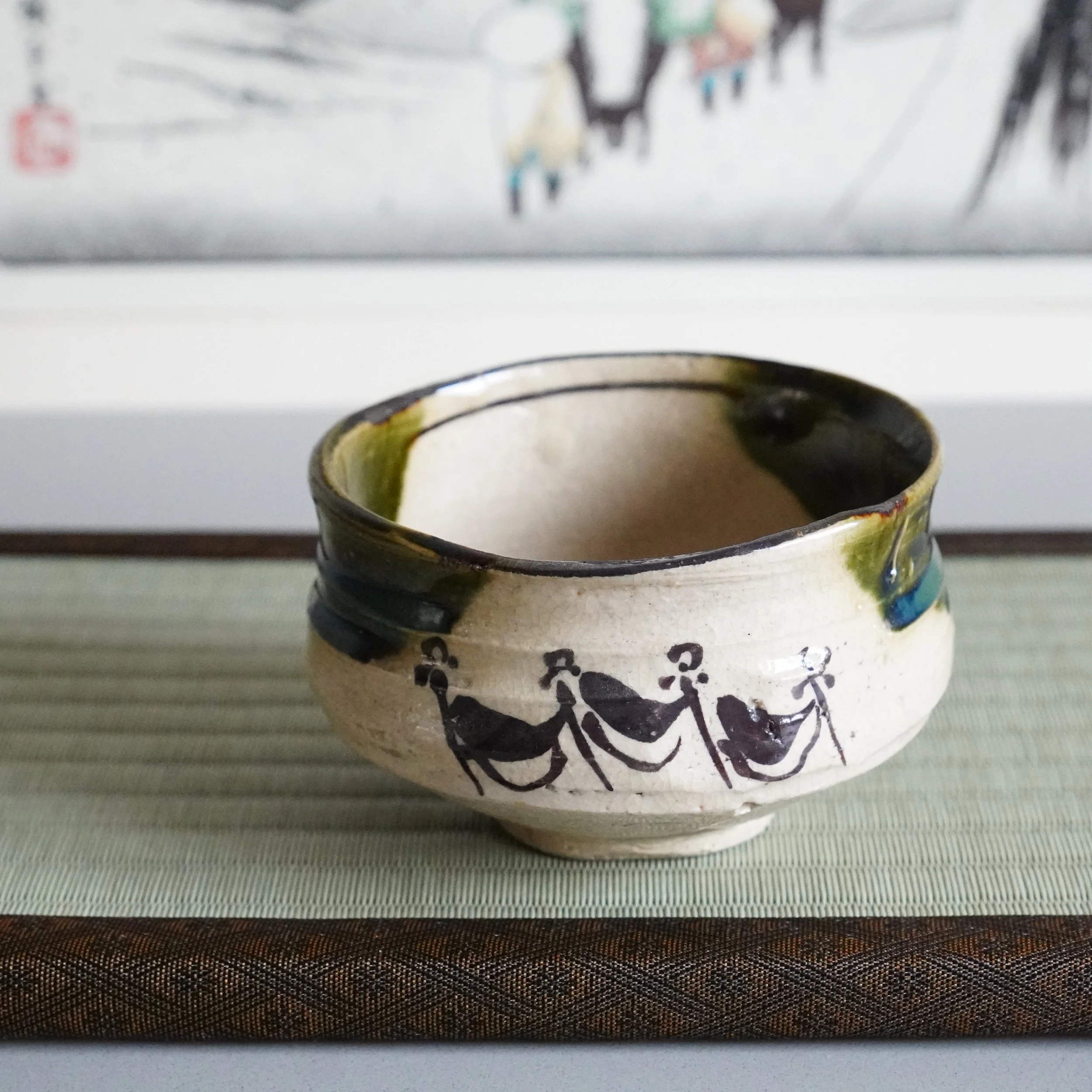 Ao-Oribe matcha bowl front view cream stoneware body with iron brushwork processional figures copper green glaze flowing over uneven rim Mino ware Showa period Japanese tea ceremony chawan on tatami