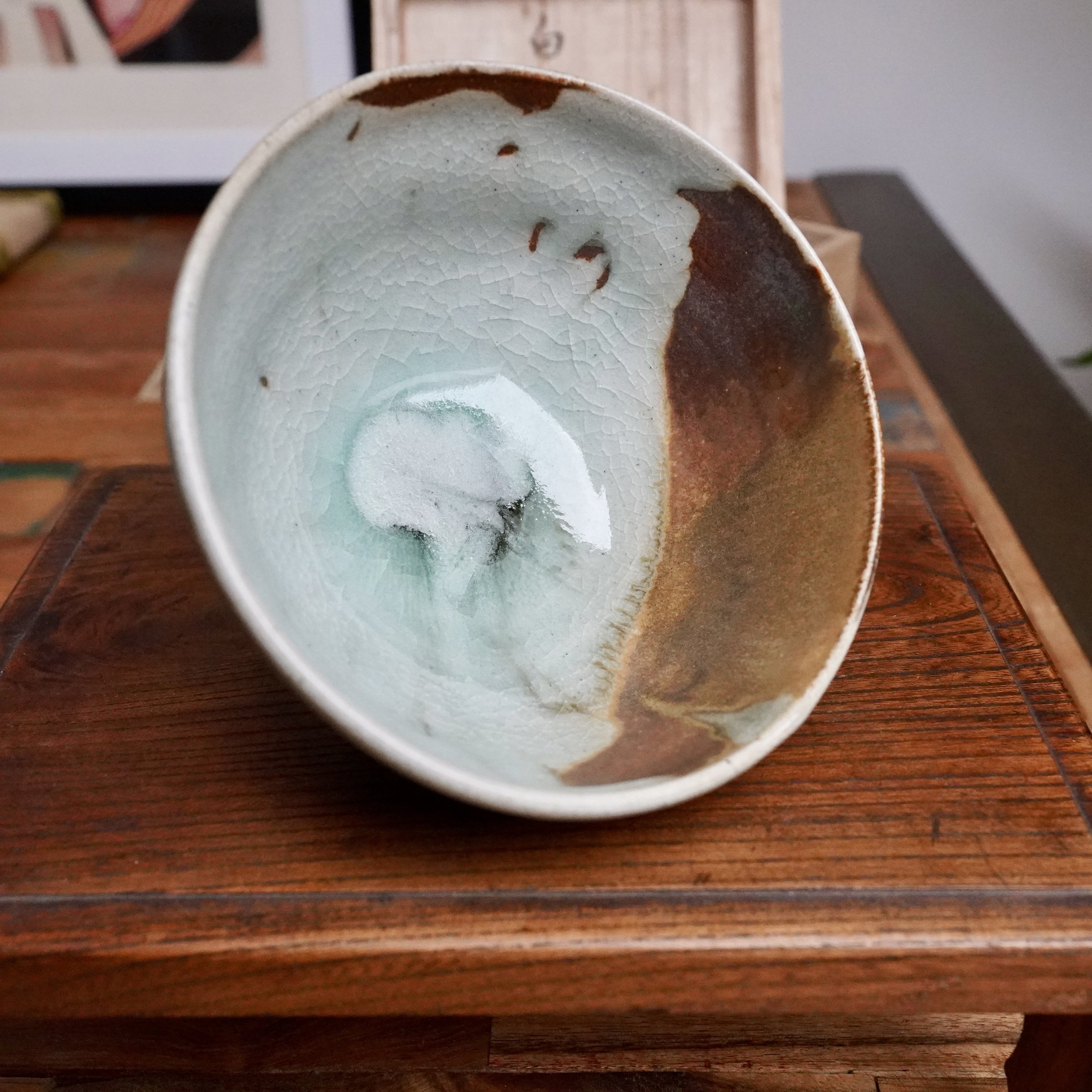 Tilted ceramic chawan, interior displaying a pale, crackled blue-grey glaze flowing into a deep rust-brown with ochre streaks, on a dark wood table.