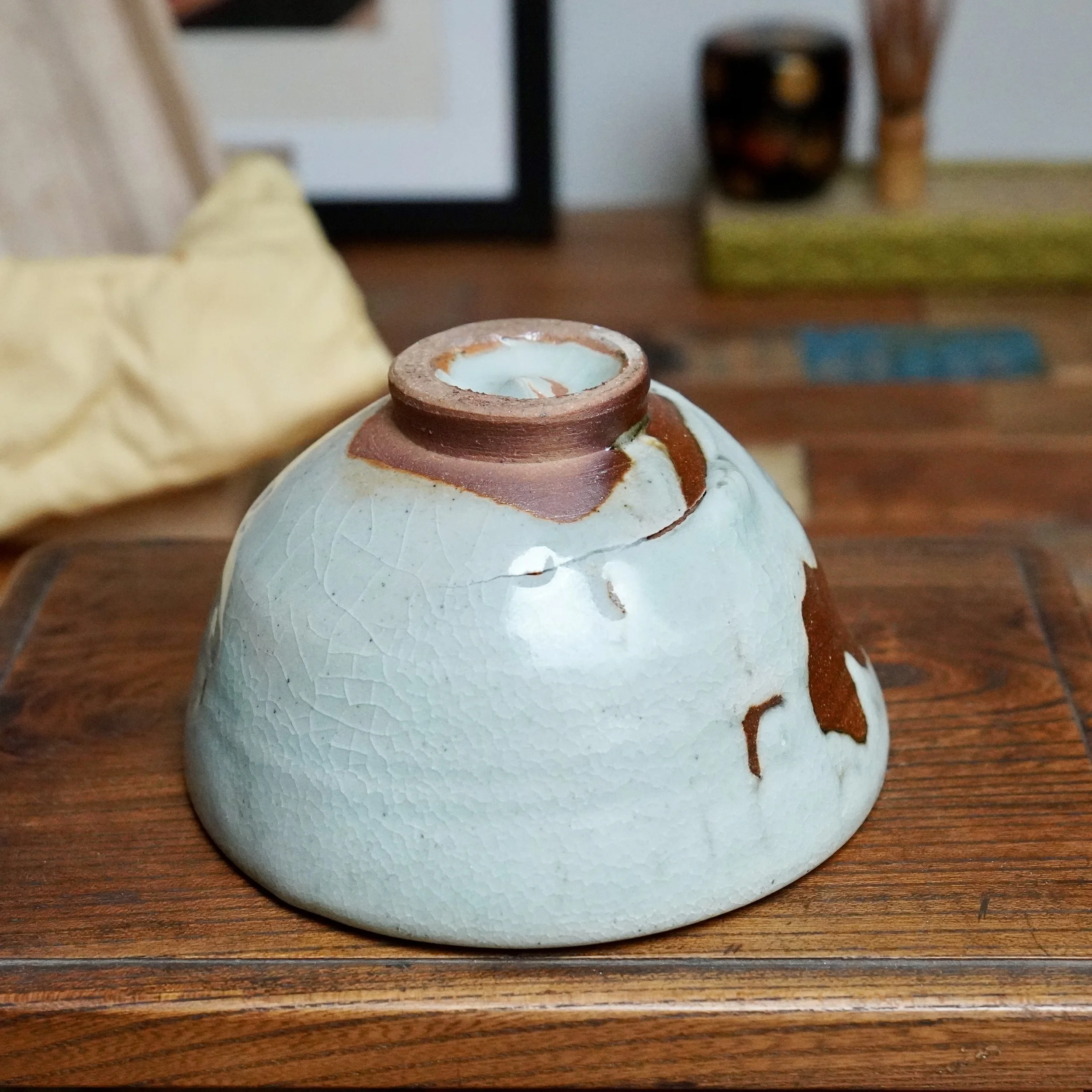 Inverted chawan with pale blue-grey crackle glaze transitioning to a deep rust-brown and ochre glaze flow on its side, revealing an unglazed red-brown foot. It sits on a wooden surface.