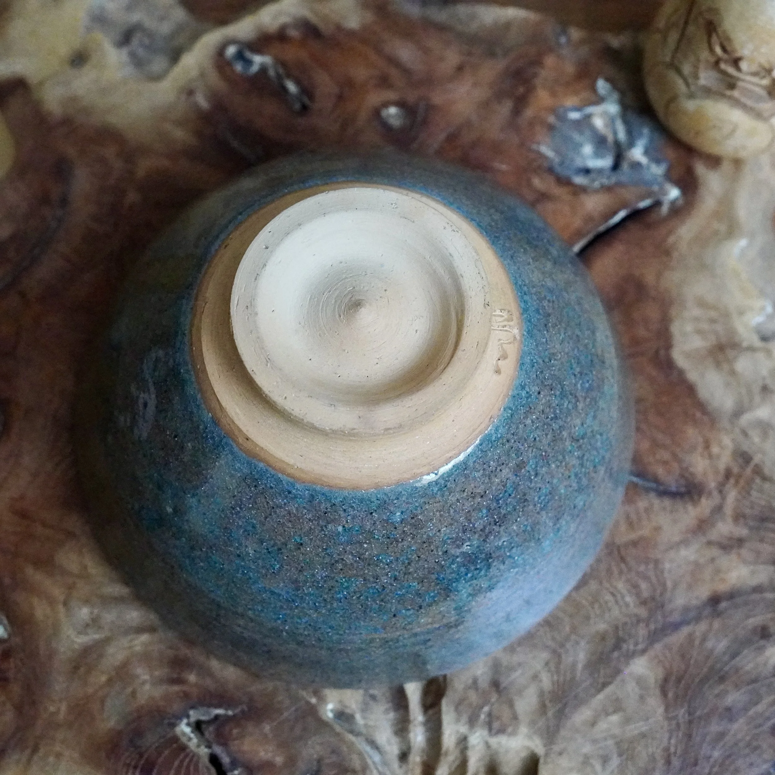 Underside of a Mizushima Jirō ceramic bowl, with a blue speckled glaze and an unglazed tan foot featuring concentric rings and a carved artist's mark, on wood.