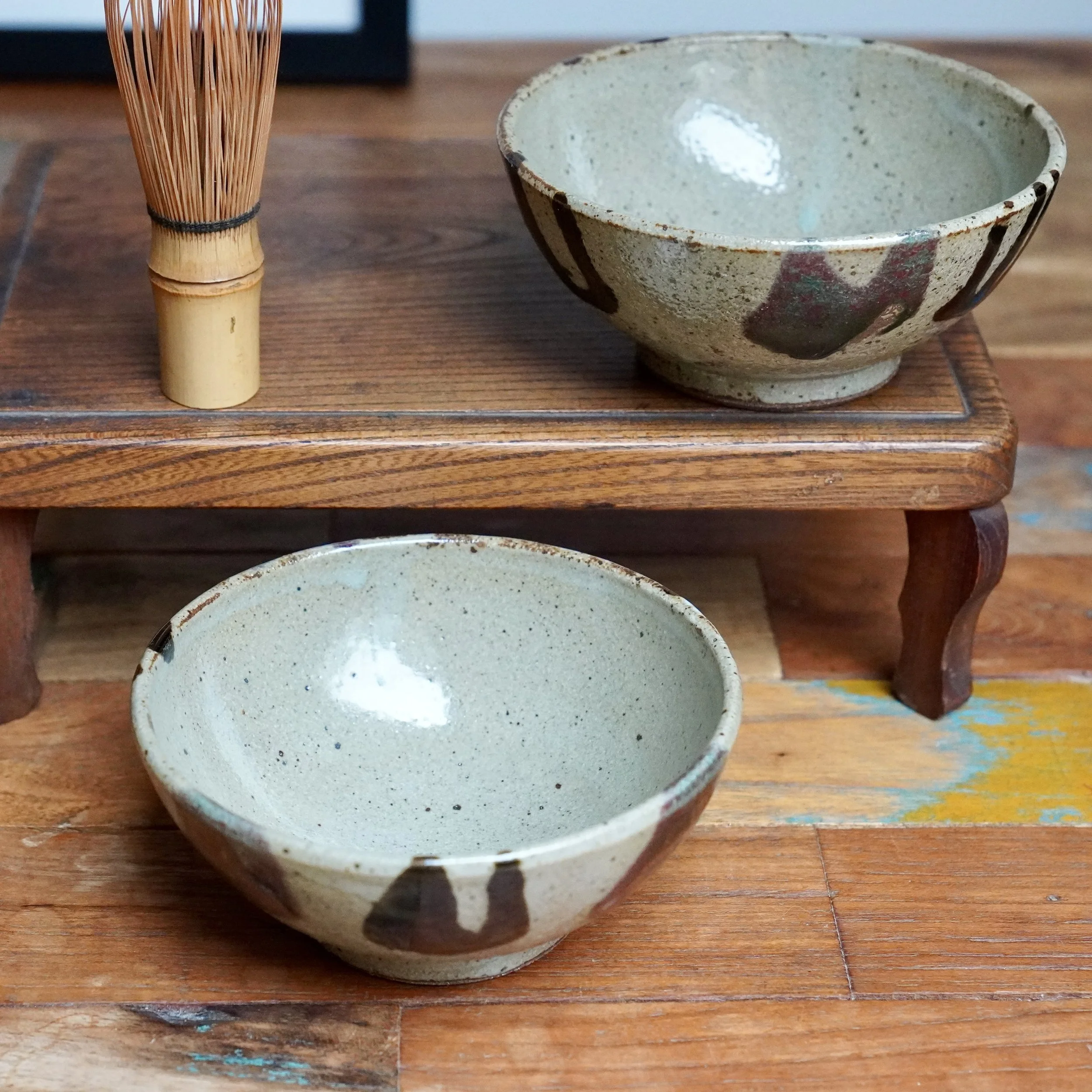 Two shallow Kuromuta-yaki bowls with speckled milky white glaze, dark brown and red strokes. One on a wooden stand with a bamboo whisk, the other on a distressed wooden floor.