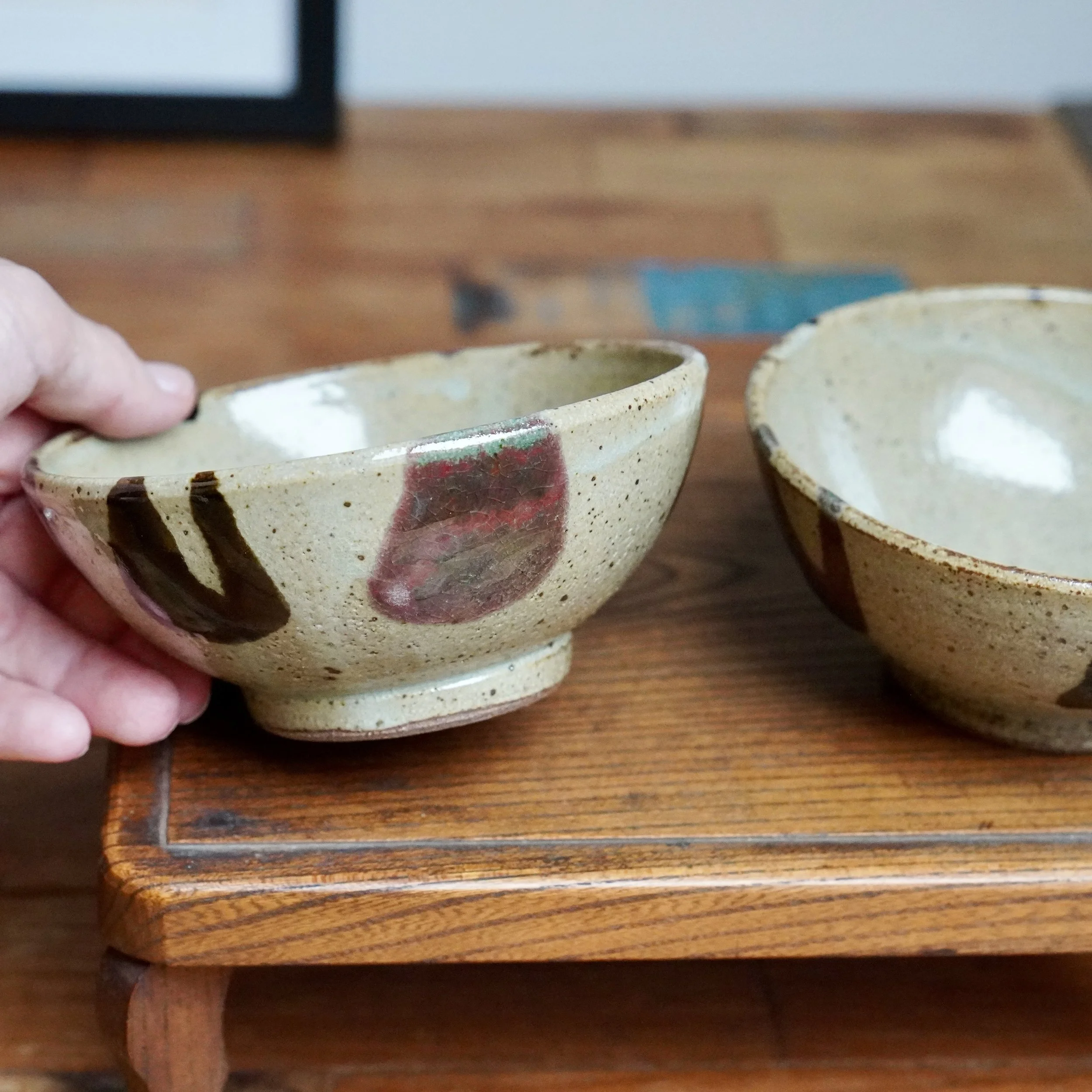 Hand holds one of two shallow, speckled ceramic tea bowls by Maruta Nobumasa, with dark brushstrokes, wine red patches, and milky glazes, on a wooden stand.