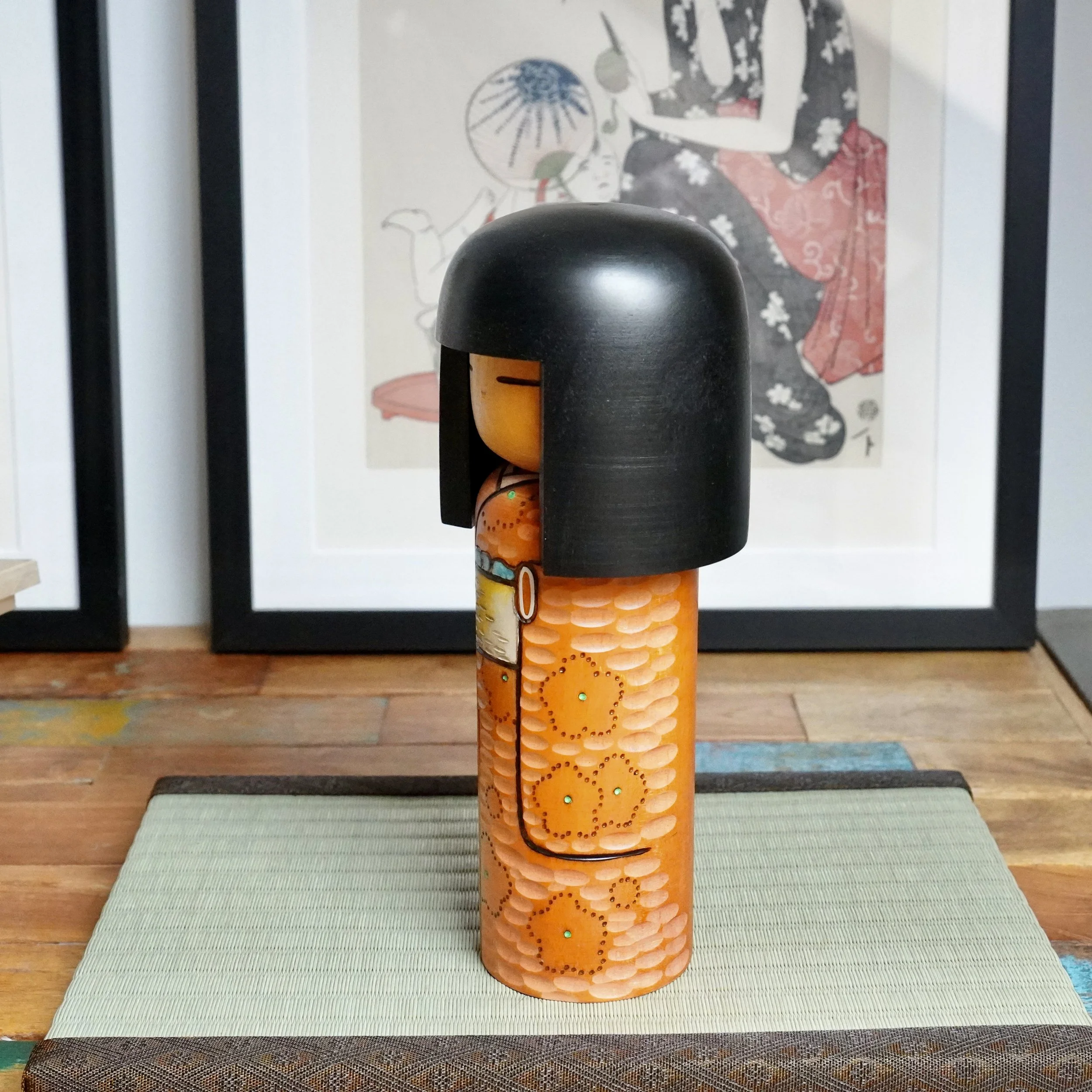 Side view of a sōsaku kokeshi by Kisaku, with black bob hair and an orange carved body detailed with painted ume blossoms, each with a green point. It stands on a light green tatami mat.