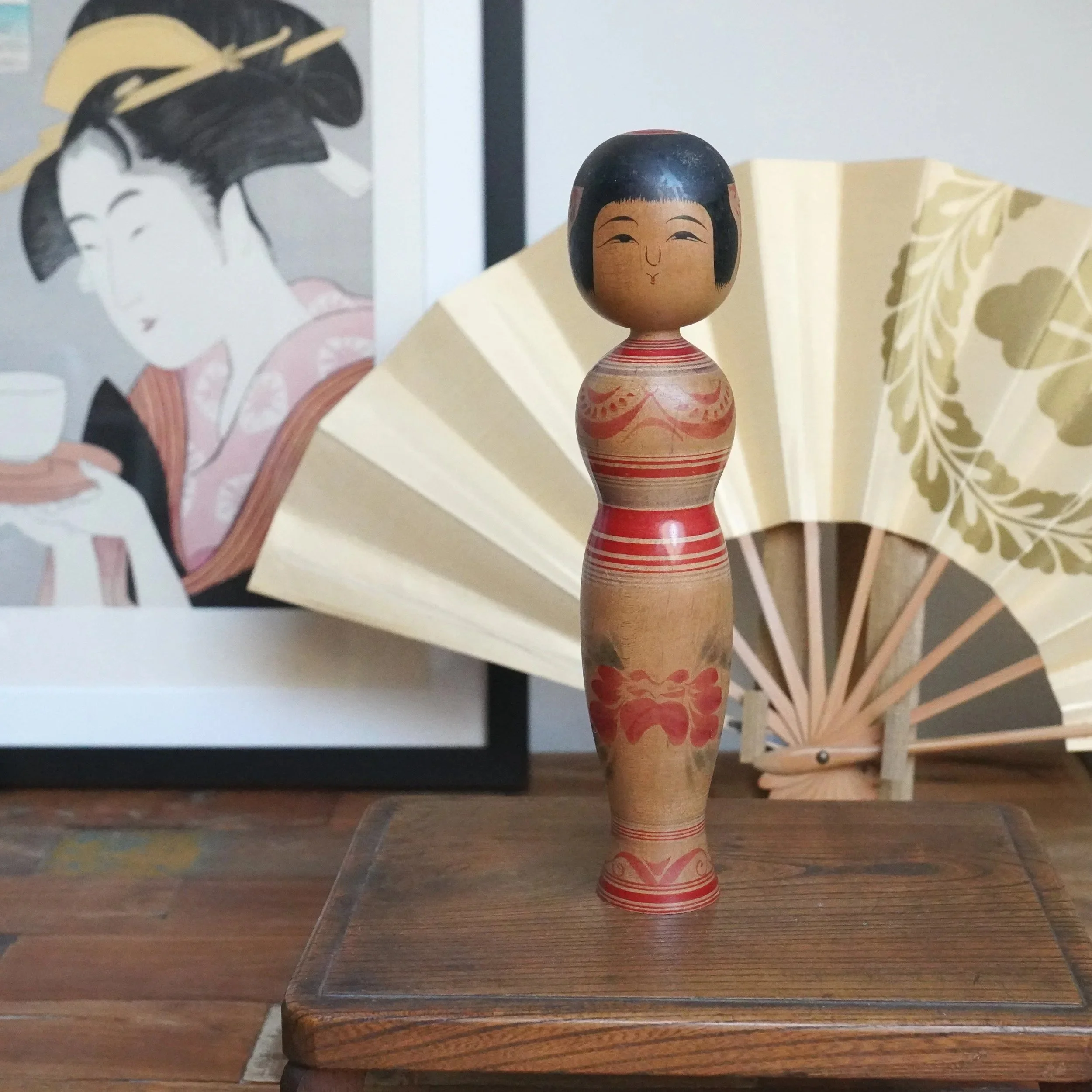 Tsugaru kokeshi with black hair, smiling face, and hourglass wood body, decorated with red stripes and a large peony. On a wooden surface, a framed print and a fan stand in the background.