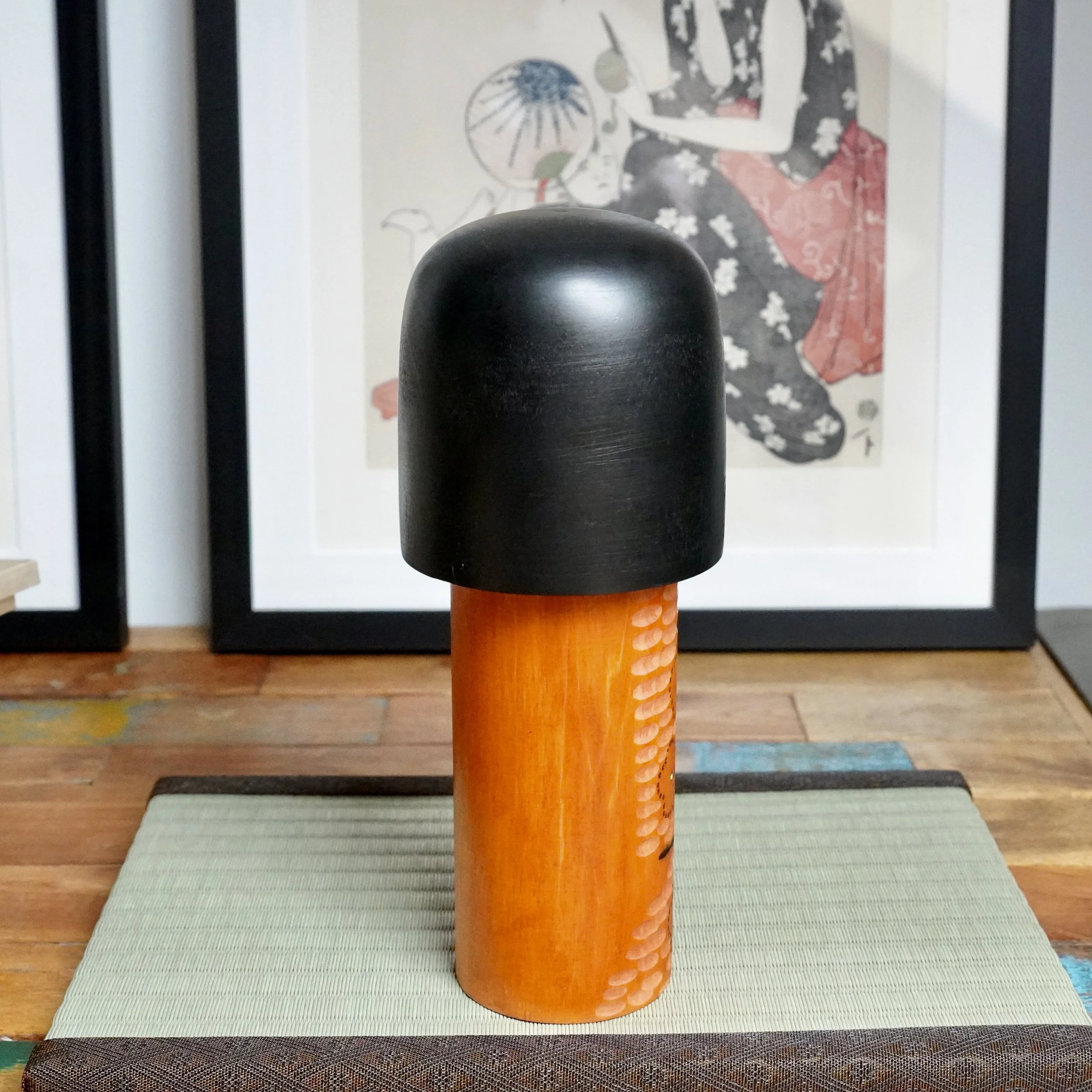 Kisaku sōsaku kokeshi with a black head and orange carved body, featuring painted light-colored ume blossoms, on a light green tatami mat.