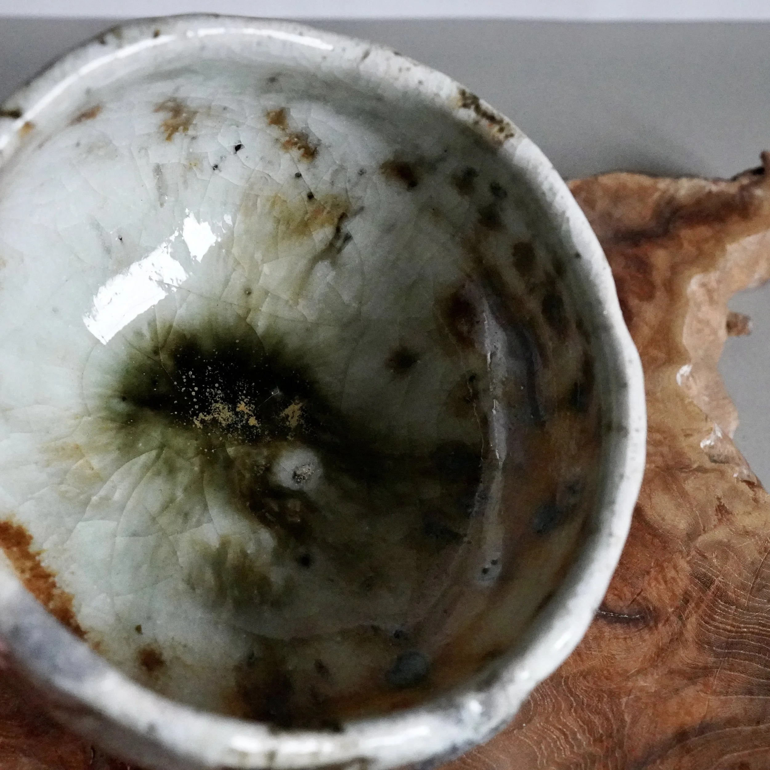 Deguchi Eiji chawan, angled close-up view of its interior with glossy crackled white glaze, deep moss green and gold pooling in the center, on rustic wood.